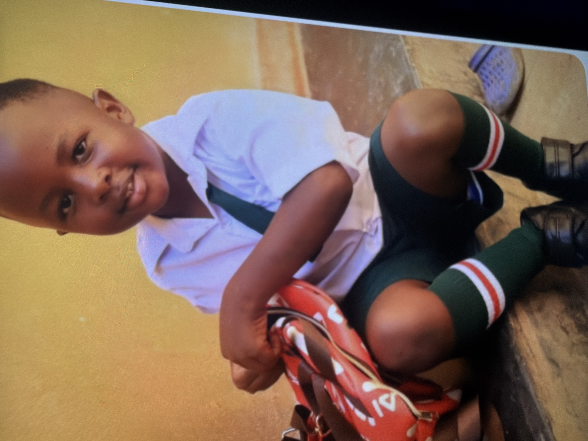 A young Black child, appearing to be of preschool age, is seated on a step, looking towards the camera with a slight smile. The child is wearing a white short-sleeved collared shirt with a dark green tie, dark green shorts, and dark green socks with red and white stripes near the top. The child's hands are resting on a red and black backpack adorned with white patterns, possibly hearts or letters. A light blue Croc-style shoe is visible on the ground to the right of the child. The background consists of a textured yellow wall. The overall lighting is warm and natural, suggesting an outdoor setting. The image appears to be a photograph taken indoors or in a covered outdoor area due to the close cropping and lighting.