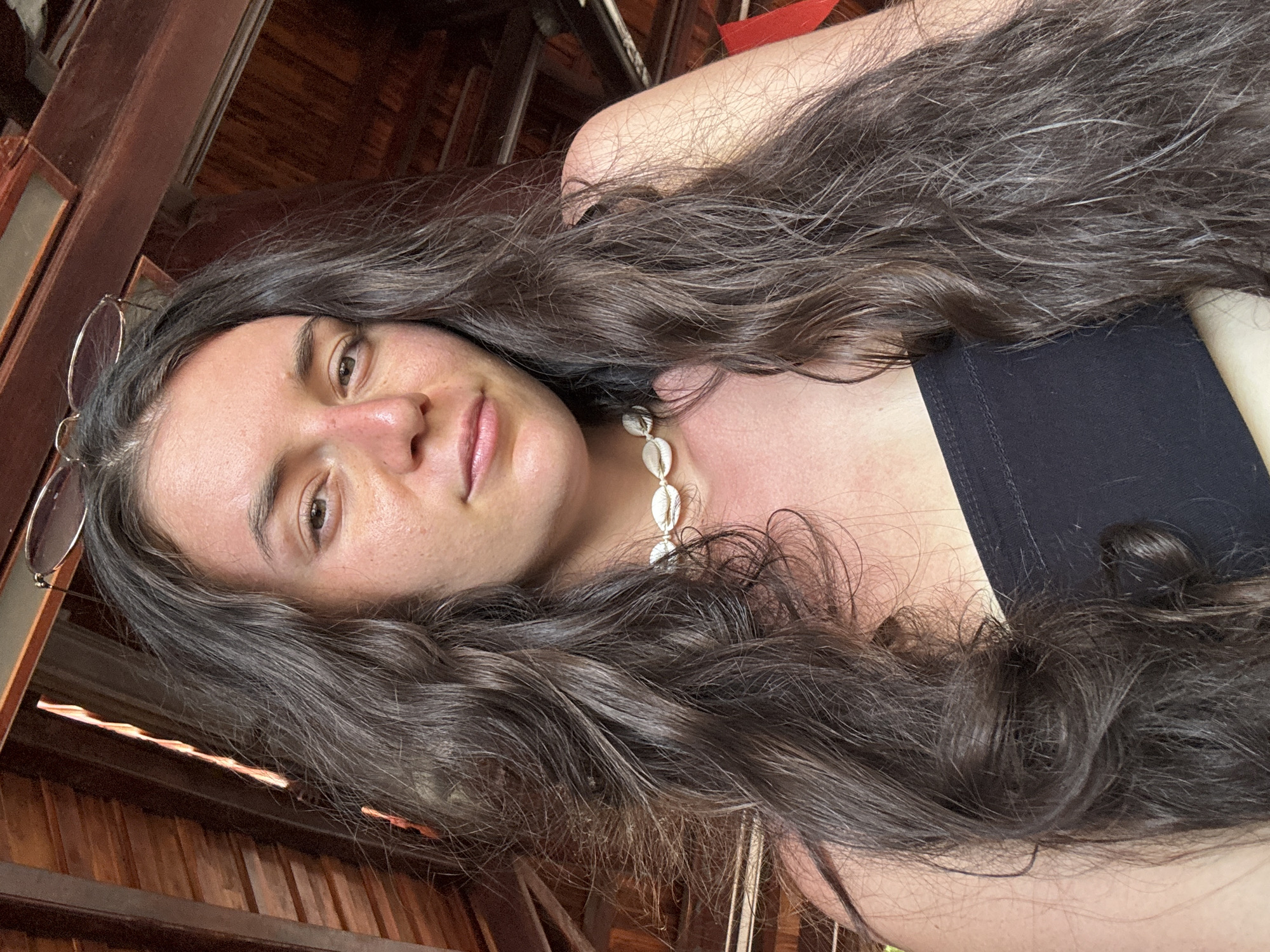 A woman with long, wavy brown hair is depicted from a low angle, looking down at the camera. She is wearing a black top and a seashell necklace. Her glasses rest on a wooden structure behind her. The background consists of wooden beams, suggesting an interior space, possibly in the city of Mũi Né, Vietnam. The natural skin texture of her face is visible.