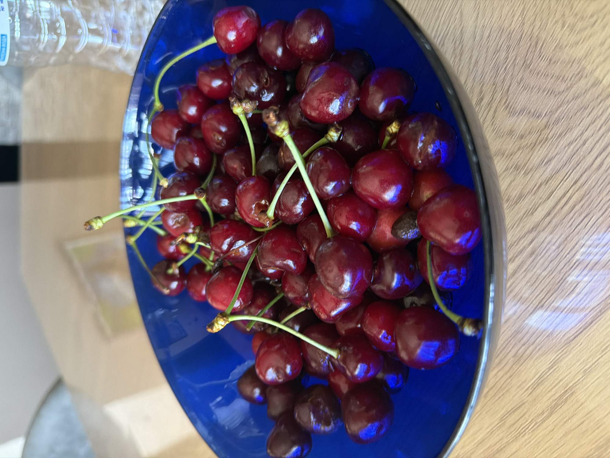 This is a top-down, close-up, outdoor shot of a bright blue bowl filled with dark red cherries. The bowl is placed on a light-colored wooden surface, with some blurred background elements including a plastic bottle and what appears to be a wall with a framed picture. The cherries are fresh and appear ripe, with their stems still attached. Some are glossy and reflecting the light, suggesting they may be wet or recently washed. The scene is lit by natural light, likely from a window, creating highlights and shadows on the fruit. There is no visible text in the image. The setting appears to be a home kitchen or dining area. The overall mood is one of freshness and simple abundance.