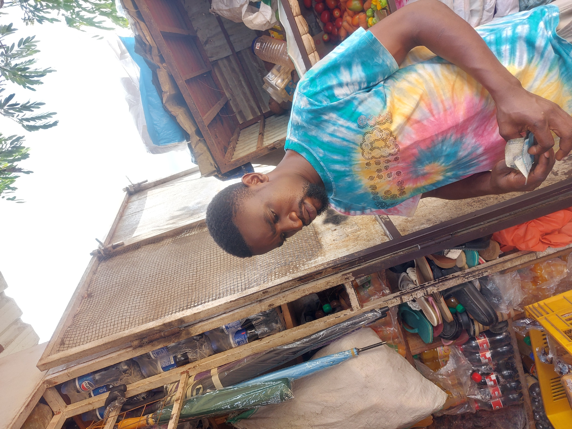A man stands in front of his kiosk in Dar es-Salaam, Tanzania, counting money. He is wearing a tie-dye t-shirt with jeweled embellishments at the shoulder. The kiosk, made of wood and metal, has a hinged, wire-mesh front panel that is open. A variety of items including shoes, umbrellas, bottled drinks, and fruits are stocked within. An umbrella is draped over a large white sack on the kiosk floor.