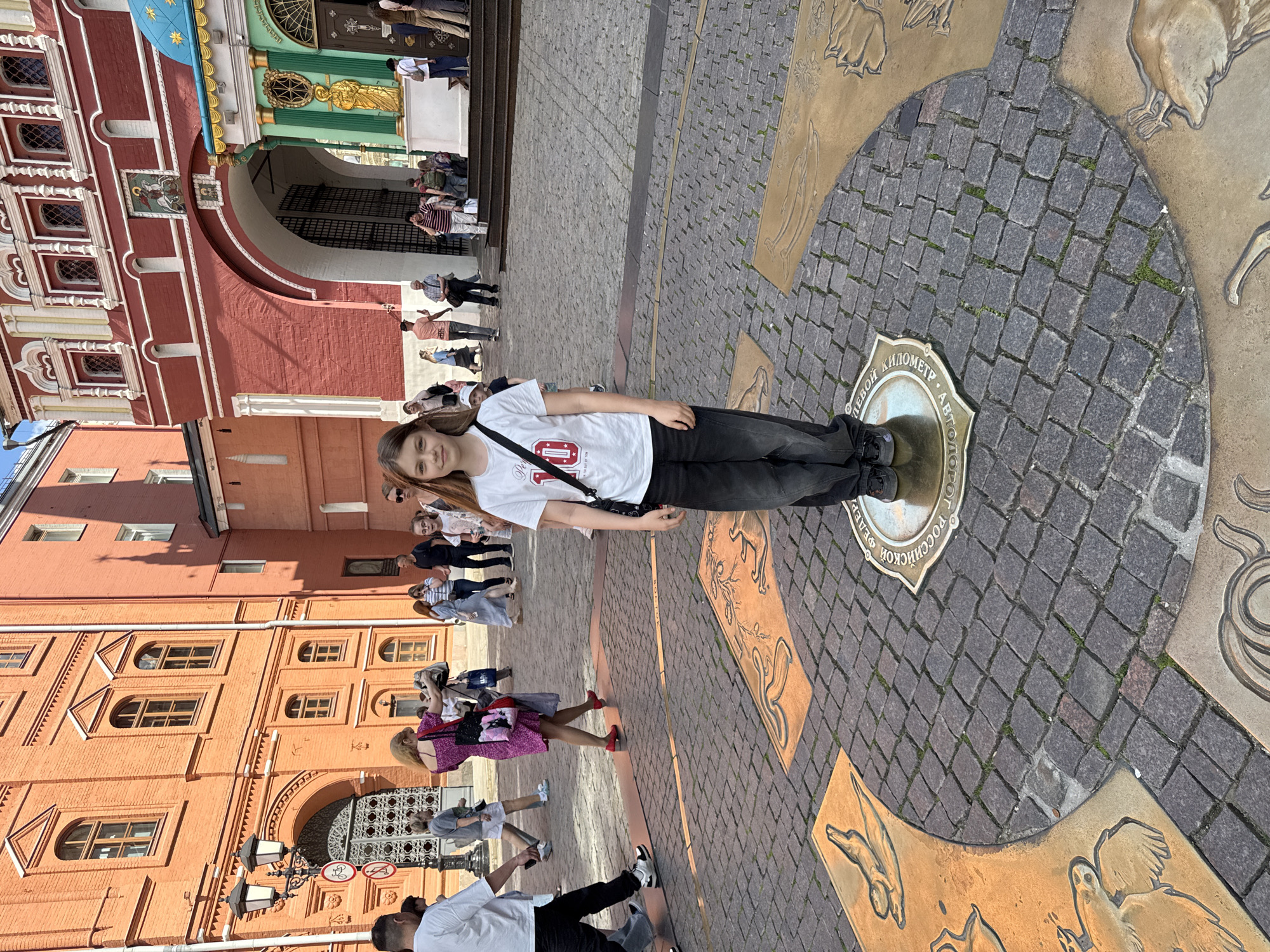 A young girl with long brown hair, wearing a white t-shirt, black pants, and a black shoulder bag, stands center-frame on a circular bronze medallion embedded in the cobblestone pavement. She faces the camera with a slight smile. Her feet are positioned directly on the medallion.
The medallion features Cyrillic text around its edge, which reads: "НУЛЕВОЙ КИЛОМЕТР • АВТОДОРОГ РОССИЙСКОЙ ФЕДЕРАЦИИ" (Zero Kilometer • Of Roads of the Russian Federation). The cobblestone ground is also adorned with larger, decorative bronze plates depicting various animal figures and mythical creatures, forming a patterned walkway.
In the background, to the left, is a distinctive red brick building with white architectural details, arched windows, and a green and gold entrance archway. A golden figure is visible above this arch. This structure appears to be part of the State Historical Museum or the Iberian Gate complex. Further to the left, another reddish-orange building with multiple windows and intricate brickwork is visible.
Several other people are present in the scene, walking or standing in the background, indicating a public, tourist-frequented area. The lighting suggests it is a bright, sunny day, with clear shadows cast on the ground. The overall atmosphere is that of a bustling urban square. The text "07" and "NEW YORK" are faintly visible on the girl's t-shirt. The setting is Moscow, Russia, specifically at the Zero Kilometer marker, often visited by tourists.