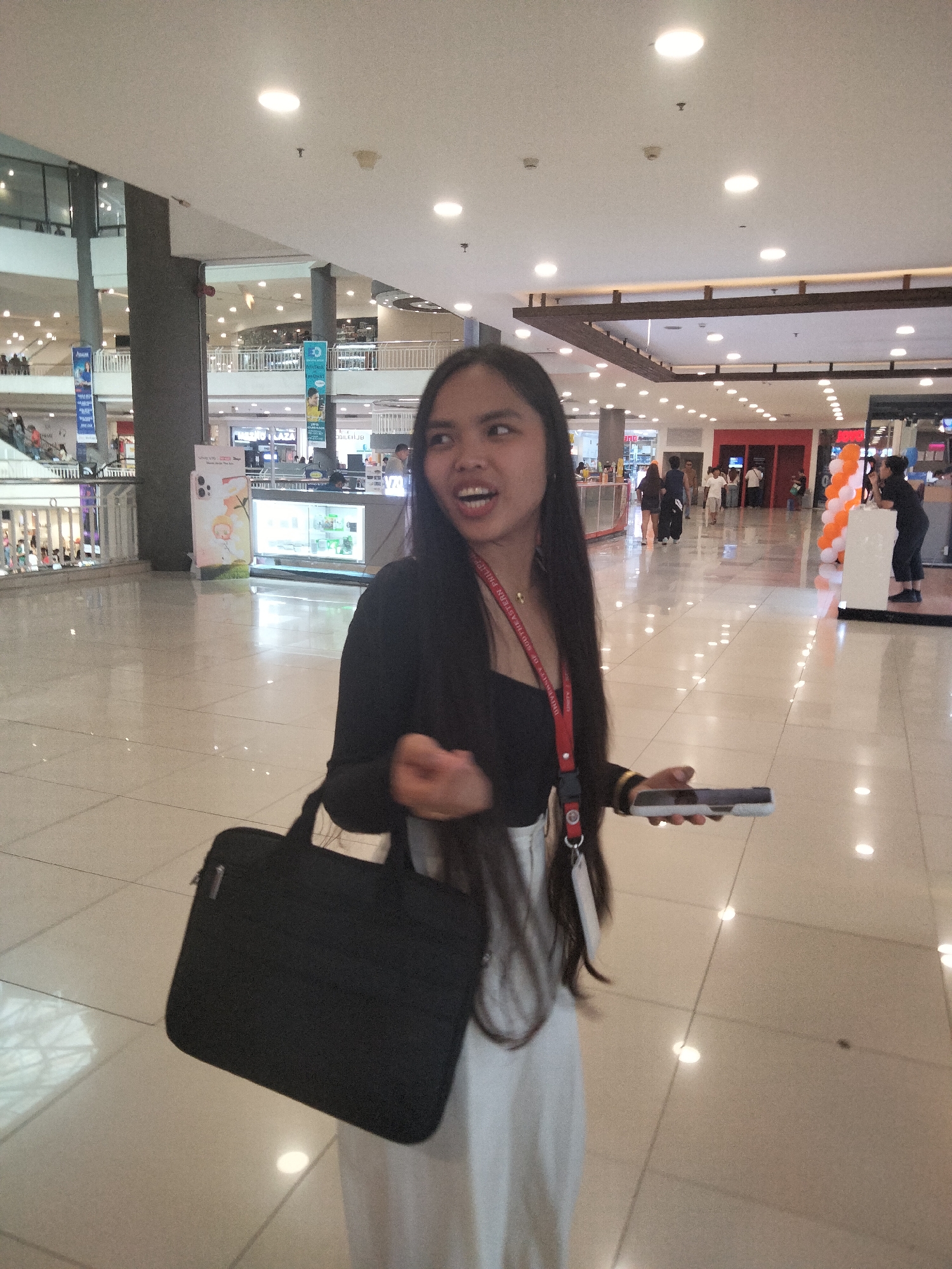 A young woman stands in a brightly lit, multi-level shopping mall in Davao City, Philippines. She has long, dark hair and is wearing a black top, white pants, and a red lanyard. She is holding a black laptop bag on her left side and a smartphone in her right hand. Her mouth is open as if she is speaking or reacting to something off-camera. In the background, there are various retail stores, displays, and other shoppers. The floor is tiled and reflective.