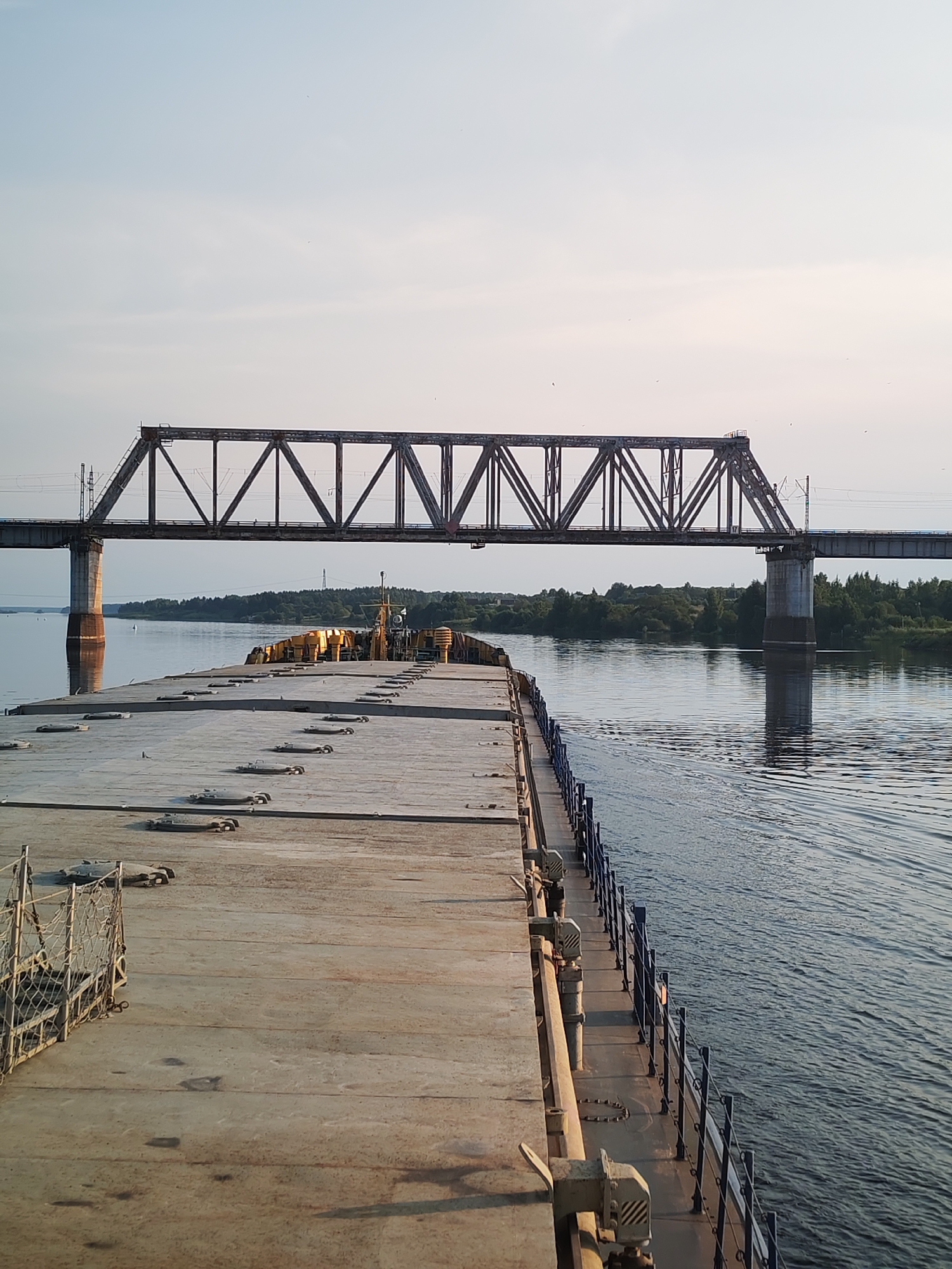 A wide, calm river flows under a metal truss railway bridge. The perspective is from the deck of a barge, looking forward towards the bridge. The barge's deck is concrete, weathered and marked with various metal fittings and covers. On the left side of the barge's deck, a metal cage-like structure with netting is visible. A dark railing runs along the right side of the barge's deck, bordering the water. The river is still, with gentle ripples, and reflects the light blue sky and the bridge. On the far bank of the river, a dense line of green trees forms the horizon. The sky is a pale, clear blue, with a few wispy clouds. The scene appears to be set in Tarkanovo, Russia, based on the context provided. There are no people visible in the image.