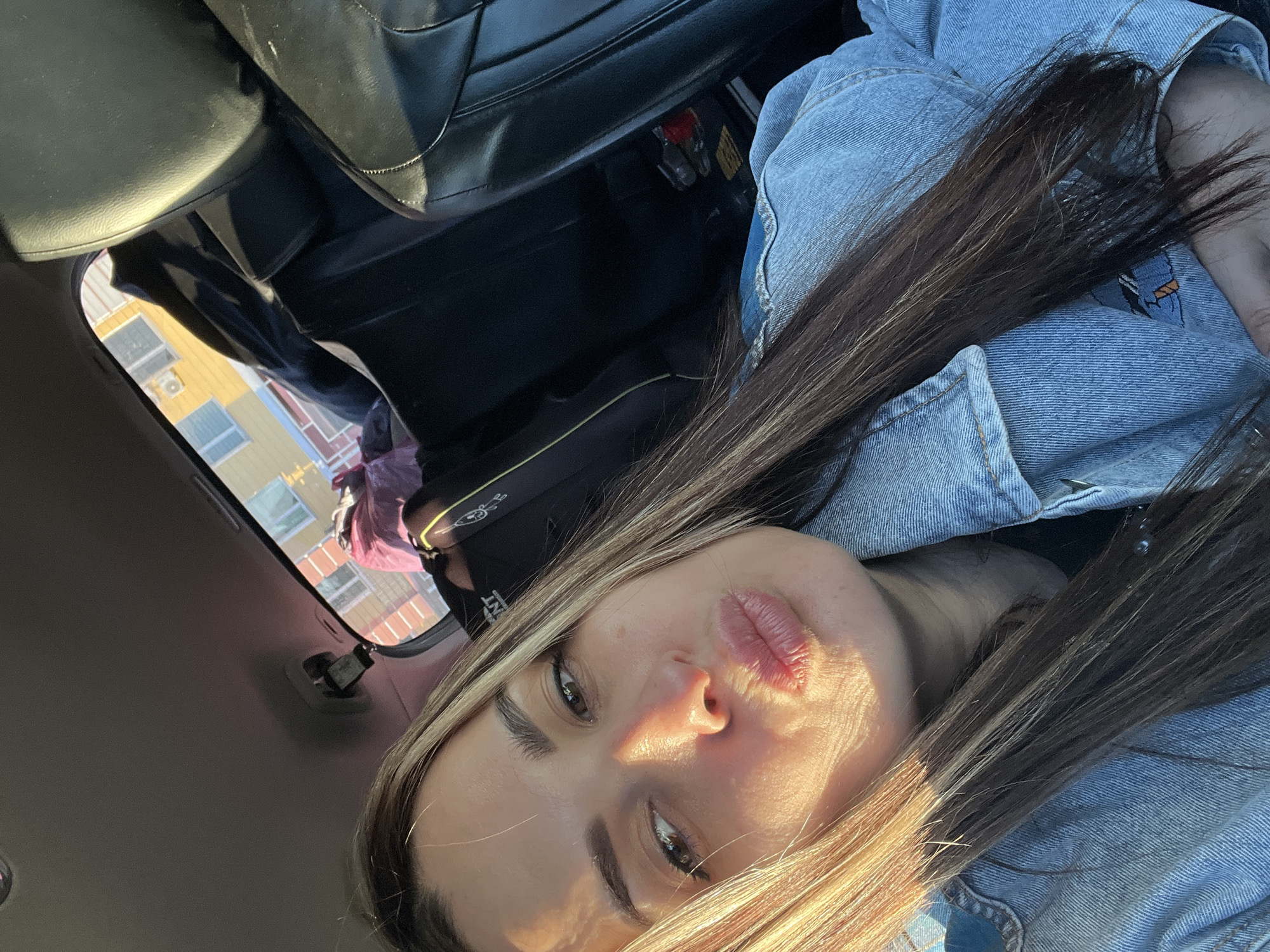 A selfie of a person with long brown hair wearing a denim jacket inside a vehicle. The person is looking directly at the camera with a pouting expression. Light is shining on their face. The background shows parts of the vehicle's interior, including a black leather seat and a view through the window of a yellow building. The location in the city is Unknown, Unknown.