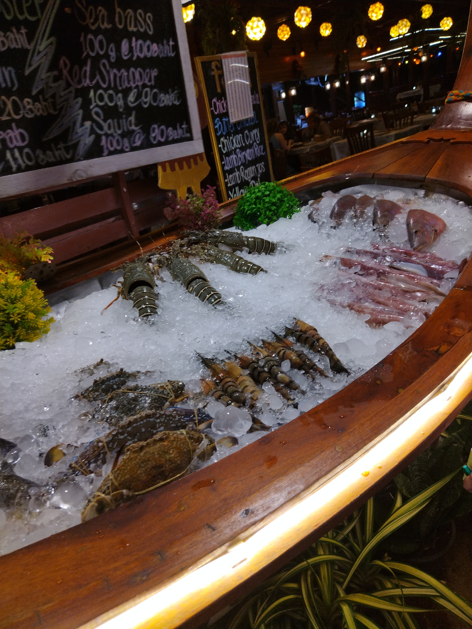The image depicts a seafood display in a restaurant setting in Patong, Thailand. The foreground features a wooden boat filled with an assortment of fresh seafood on ice, including lobsters, crabs, squid, and fish. The seafood is arranged artistically, showcasing its freshness. In the background, a blackboard menu displays prices for various seafood items. The restaurant appears to be an outdoor or semi-outdoor establishment, with wooden tables and chairs visible, and decorative lighting overhead. The overall atmosphere suggests a lively dining environment, possibly in the evening, given the lighting.