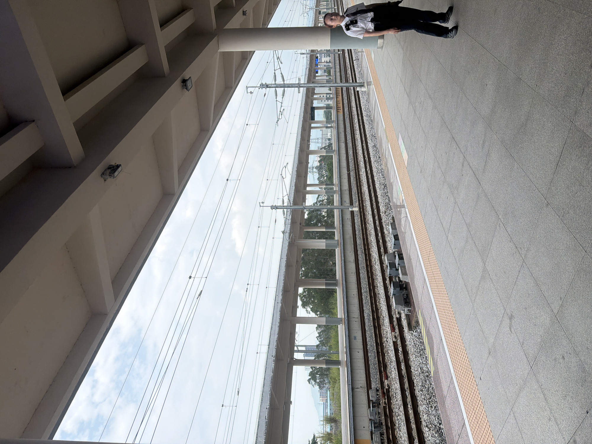 An exterior, eye-level, medium shot captures a woman in a white shirt and dark pants standing on a train platform in Jiangmen, China. She is facing to the right, away from the camera, and looking down the railway tracks. She has short, dark hair pulled back and is wearing a dark tie. The platform is paved with large gray tiles, with a yellow tactile warning strip along the edge. Railway tracks run parallel to the platform, filled with gravel and ballast. Overhead, a concrete structure with visible beams and lighting fixtures provides shade for the platform. The sky is overcast with scattered clouds. In the distance, a covered walkway supported by pillars extends along the railway line. Trees and buildings are visible beyond the tracks, suggesting an urban or semi-urban environment. The overall scene is one of quiet anticipation at a transportation hub.