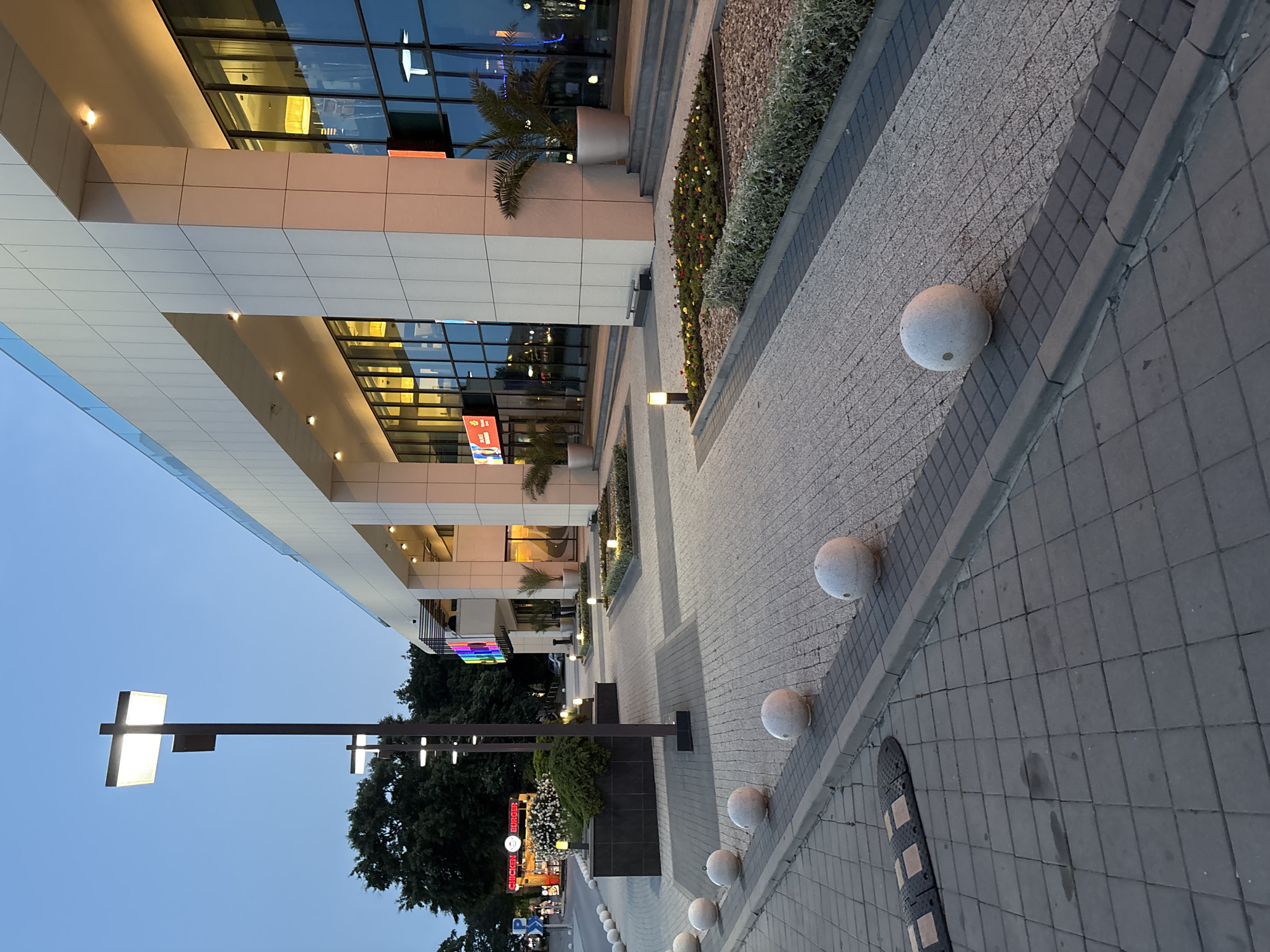 The image displays the exterior of a modern commercial complex, likely a shopping center or mall, located in Varna, Bulgaria, observed during late twilight or early evening. The sky is a clear, light blue.
In the foreground, a dark gray square-tiled sidewalk is visible on the left, featuring a black and tan speed bump. A row of at least six spherical, light-gray concrete bollards runs parallel to the sidewalk, demarcating a pedestrian area. A dark gray curb separates these elements from the central paved area, which is laid with light gray aggregate pavers bordered by darker gray blocks.
The midground features several tall, dark-colored lampposts, each fitted with two bright, rectangular white light fixtures. A prominent, large, dark gray tiled planter box filled with dense green shrubbery and containing small ground-level light fixtures is centrally positioned. To the far left, a "Chicken Burger" food stand with illuminated red neon lettering is visible, alongside a partially obscured red sign that appears to read "DALMATINE HOUSE". A few indistinct human figures are present near the food stand. Closer to the main building, several decorative flower beds containing small red, yellow, and white flowers, bordered by low green hedges and illuminated by small ground lights, are arranged along the facade. Large gray planters holding palm trees are placed at various entrance points of the building.
The background is dominated by a multi-story, modern building constructed with light-colored, possibly off-white, paneling and extensive dark-tinted glass windows. The building exhibits a design with deep, recessed sections forming sheltered entrance areas or outdoor terraces. The interior spaces behind the glass are warmly lit, and the glass surfaces reflect ambient light. A glass balustrade is visible along the upper edge of the building. A multi-colored sign, possibly displaying the word "RAINBOW," is visible within one of the building's recessed architectural features. The entire scene is brightly illuminated by artificial lighting from streetlights, interior building lights, and landscape features, contributing to a clean and contemporary urban ambiance.