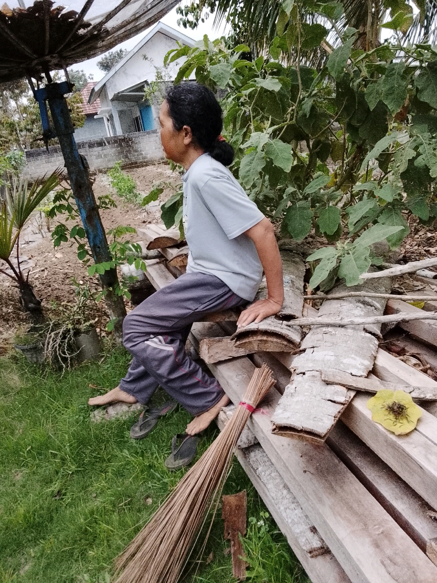 An individual sits on a stack of lumber outside a residence, facing away from the camera. The person is wearing a light blue t-shirt and dark gray pants with red accents. Their left leg is bent at the knee, with their foot resting on the ground. Their right leg is extended. A broom made of dried coconut fronds lies on the ground next to the lumber pile. A yellow leaf with dark spots is positioned on one of the planks. In the background, a white house with blue shutters is visible behind a low brick wall and a fence. Lush green foliage and trees surround the scene. The ground is covered in grass and loose soil. The scene appears to be outdoors in a residential area. The location is likely Padang Dalom, Indonesia, given the architectural style of the house and the surrounding vegetation.