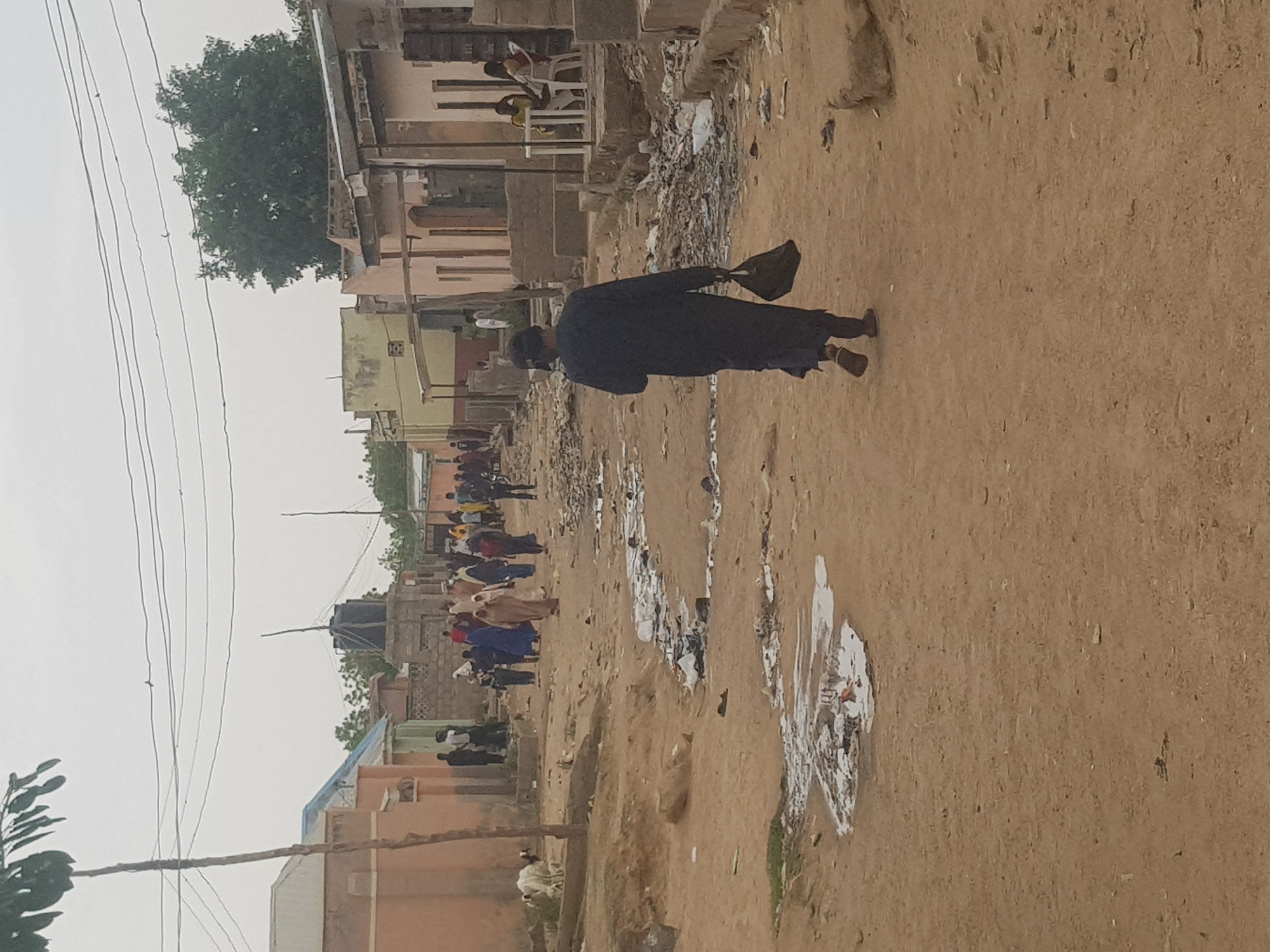 The image depicts a street scene in what is identified as Dabawa, Nigeria. A person dressed in dark clothing is walking along a dirt road. There is trash and debris scattered along the roadside. In the background, a group of people are walking or standing in the distance. Buildings and vegetation are visible on either side of the road, and electrical wires are strung overhead.