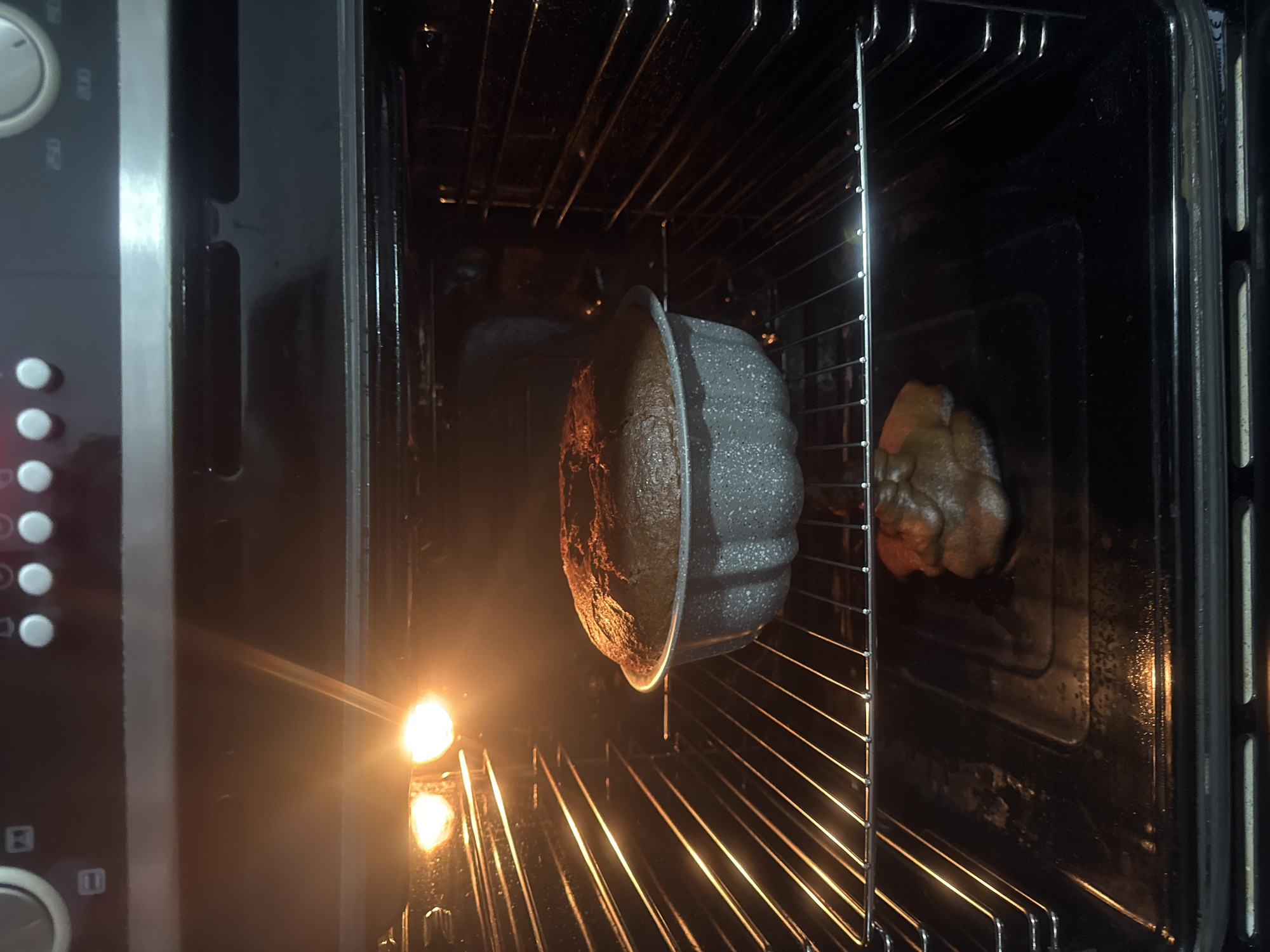 An oven's interior is illuminated by the glow of its heating element, and two baked items are visible. On the left, a cake in a gray Bundt pan sits on the oven rack, its top browned. To the right, a second baked item sits on the rack. A lit match is positioned just inside the oven door. The setting suggests an indoor kitchen environment, potentially in Tbilisi, Georgia, based on the details of the items. The scene depicts a moment in the process of baking.