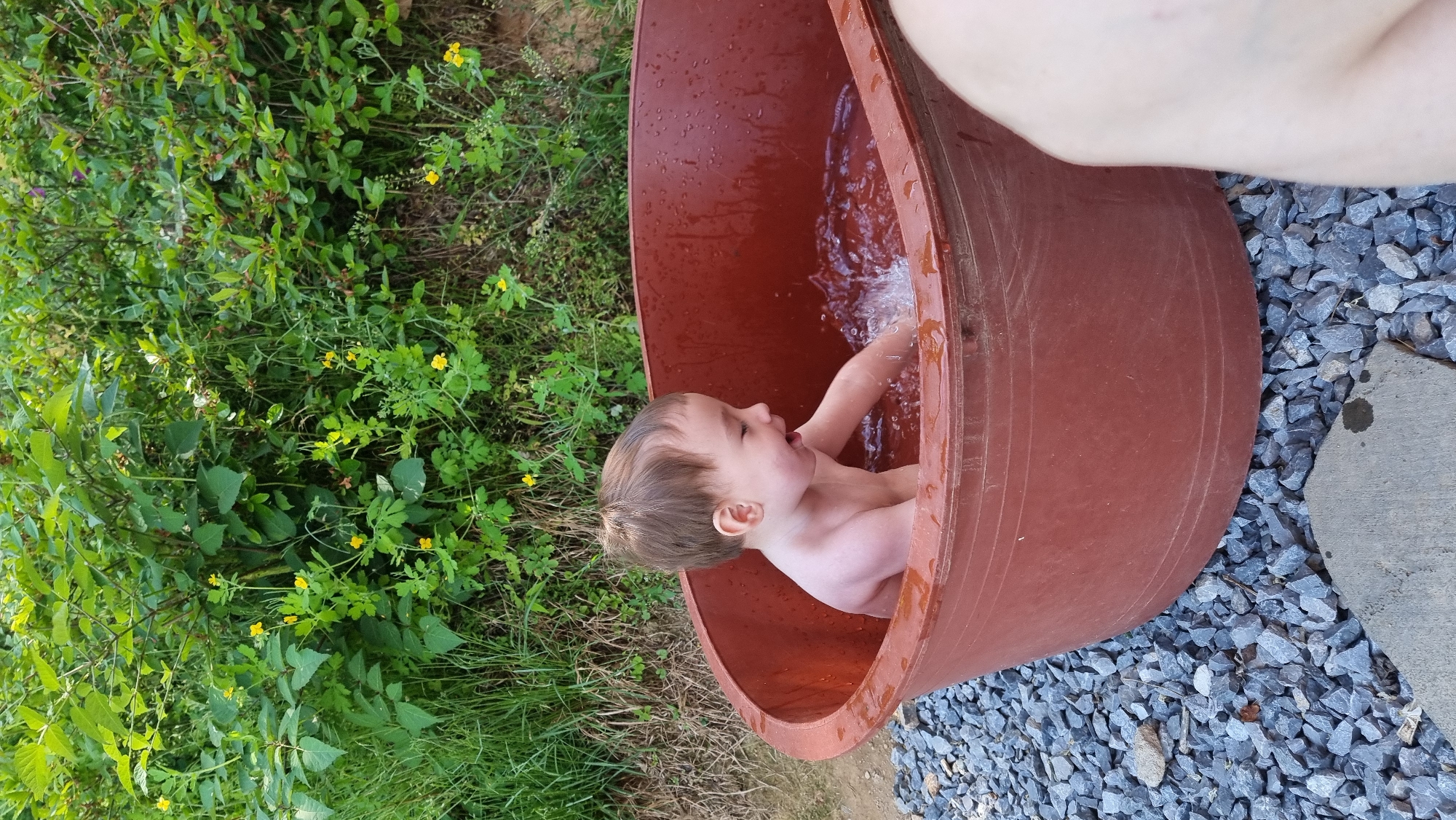 In Dunnae-myeon, South Korea, a young child is seated inside a large, reddish-brown plastic tub filled with water. The child is splashing water with their hand. The tub is positioned outdoors, near green foliage on one side and a patch of gray gravel on the other. A person's arm is visible near the top edge of the frame.