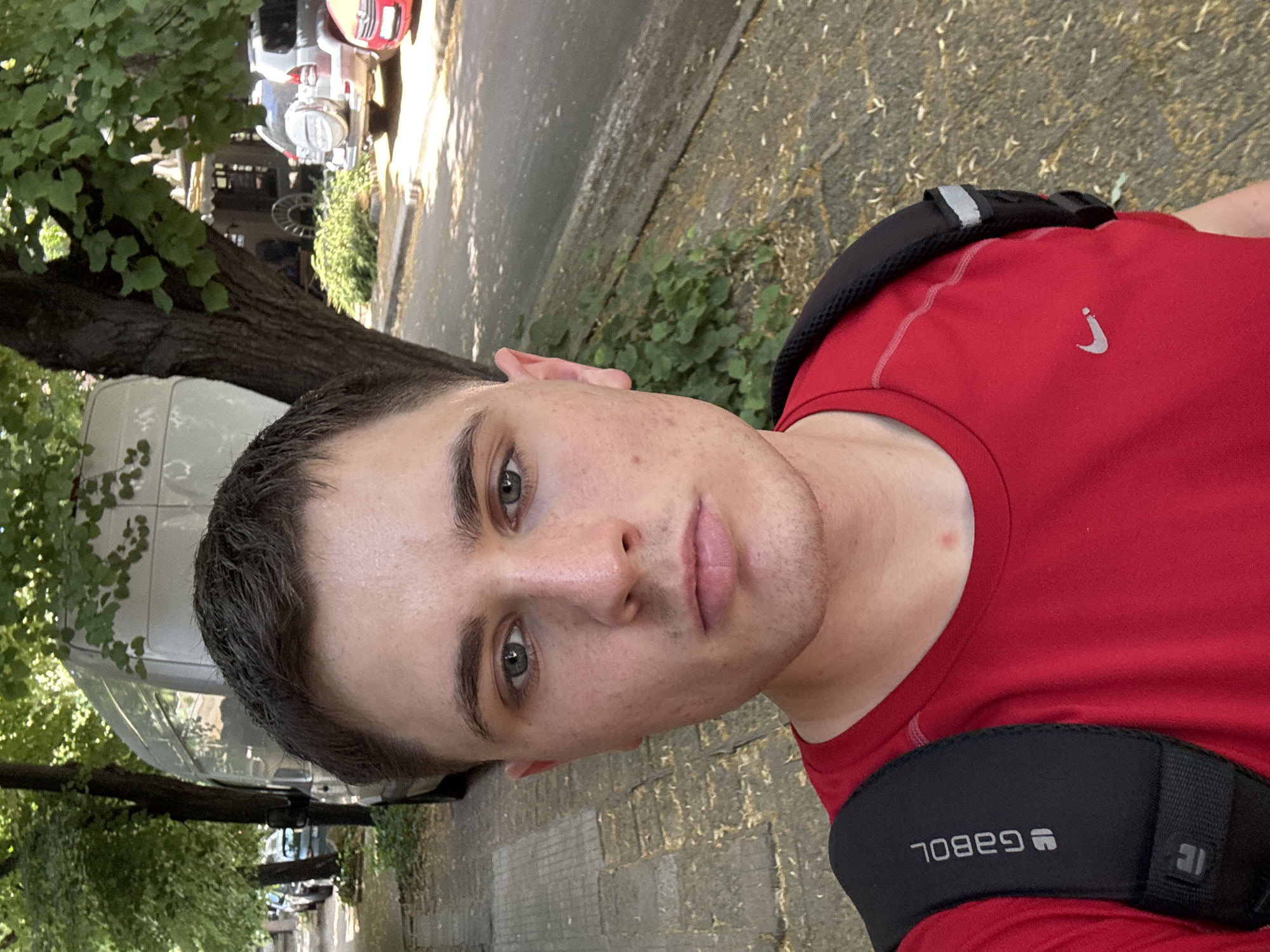 A young man with short brown hair and blue eyes, wearing a red t-shirt and a black backpack, is taking a selfie outdoors. He is standing on a sidewalk next to a paved street lined with trees. In the background, several parked cars and a white van are visible. The environment appears to be a residential or commercial street in the city of Stara Zagora, Bulgaria, given the architectural style and the presence of trees and parked vehicles common in urban Bulgarian settings. The lighting suggests it is daytime.