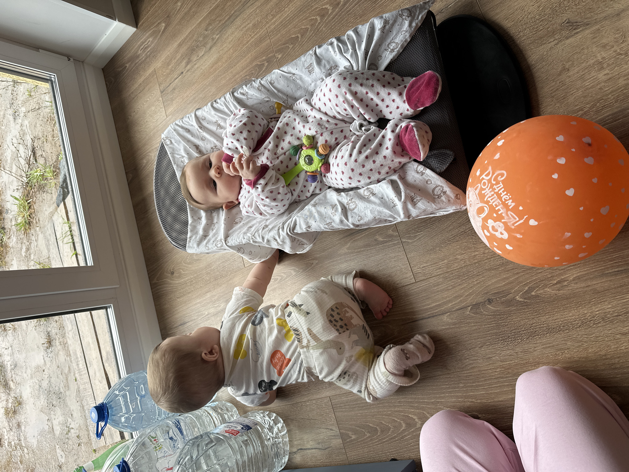 The image shows two infants on a wood floor in what appears to be an indoor setting, possibly a residence. The location may be Chaikovsky, Russia.
One infant is lying on a baby lounger wearing a patterned onesie with pink footies and is holding a toy. The other infant is on the floor in a white patterned pajama, near several large plastic water bottles. An orange balloon with writing in Cyrillic script, potentially related to a birthday, is located near the lounger. The bottom right corner of the image shows a person wearing pink pants, possibly observing the scene.