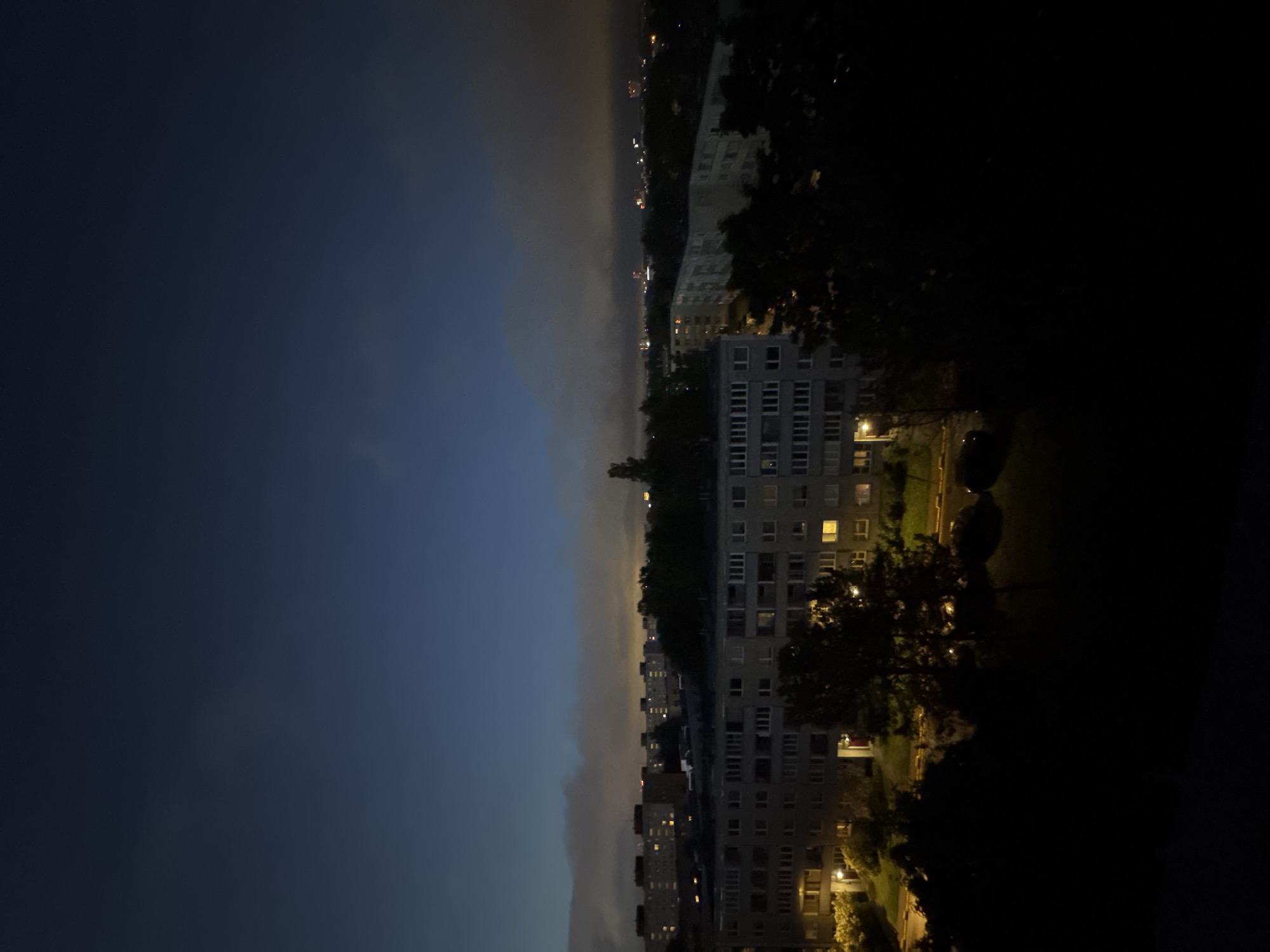 The image shows a twilight view of a residential area in Tallinn, Estonia. The sky is a deep blue with some diffused clouds and distant city lights visible on the horizon. In the foreground, silhouetted trees obscure the lower part of the scene. The middle ground is dominated by a row of multi-story apartment buildings, characterized by their flat roofs and numerous windows, many of which are illuminated with warm yellow light. A street runs in front of the buildings, with parked cars visible. The overall atmosphere is quiet and nocturnal, typical of a city at dusk or early night.