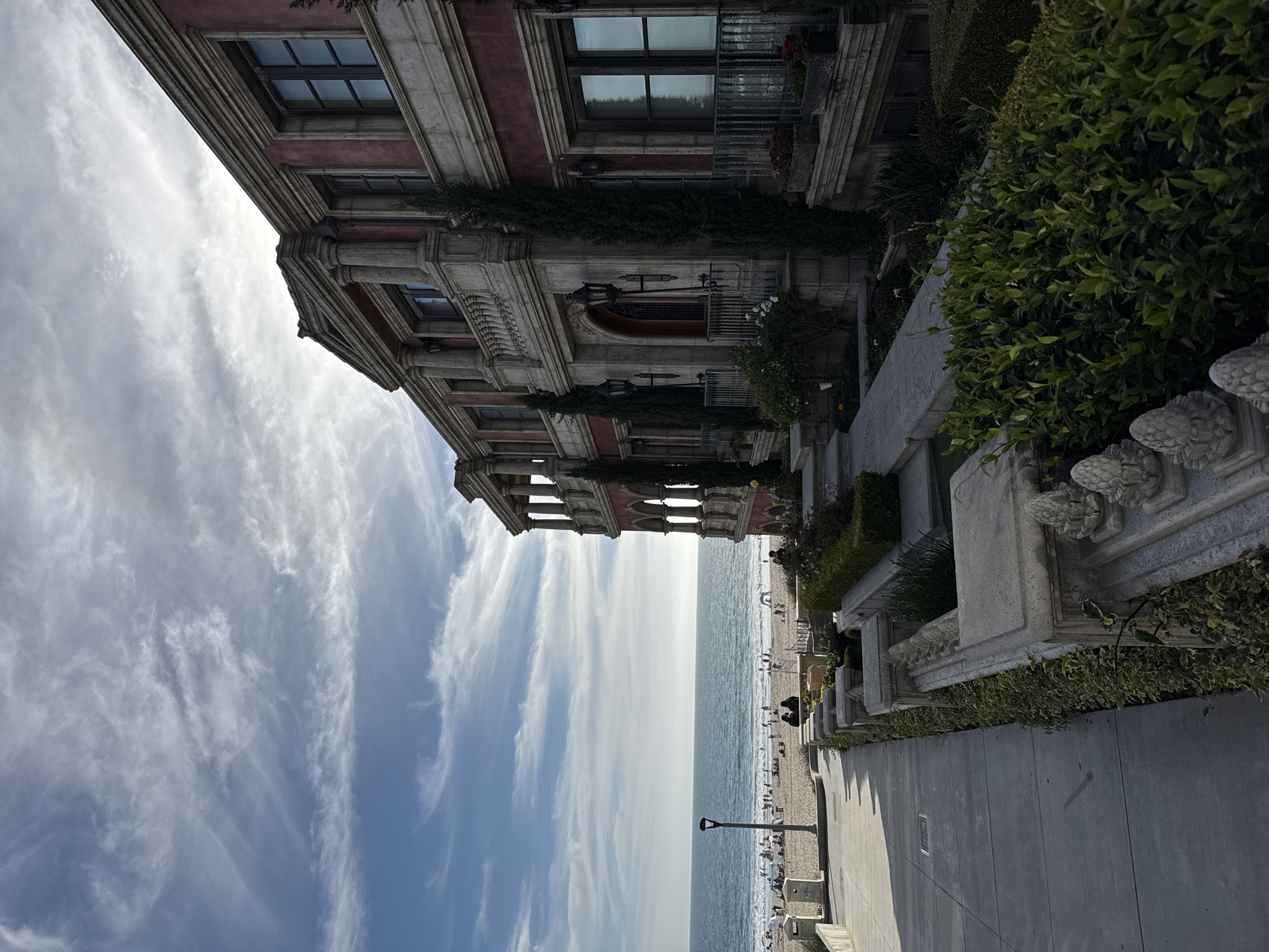 The image depicts a grand, multi-story building with red stucco walls and ornate architectural details, reminiscent of Italian Renaissance style. It is situated in Manhattan Beach, California, overlooking a sandy beach and the Pacific Ocean. The building features multiple balconies with classical balustrades, arched windows, stone columns, and decorative moldings. Cypress trees and manicured hedges are visible in the landscaping. On the beach below, numerous people are scattered, some walking, others sitting, and some are in the water. The ocean waves are gently rolling onto the shore under a sky with wispy white clouds. A lamppost stands on the promenade. The perspective is from an elevated walkway looking down towards the beach and the ocean, with the building occupying the right side of the frame.