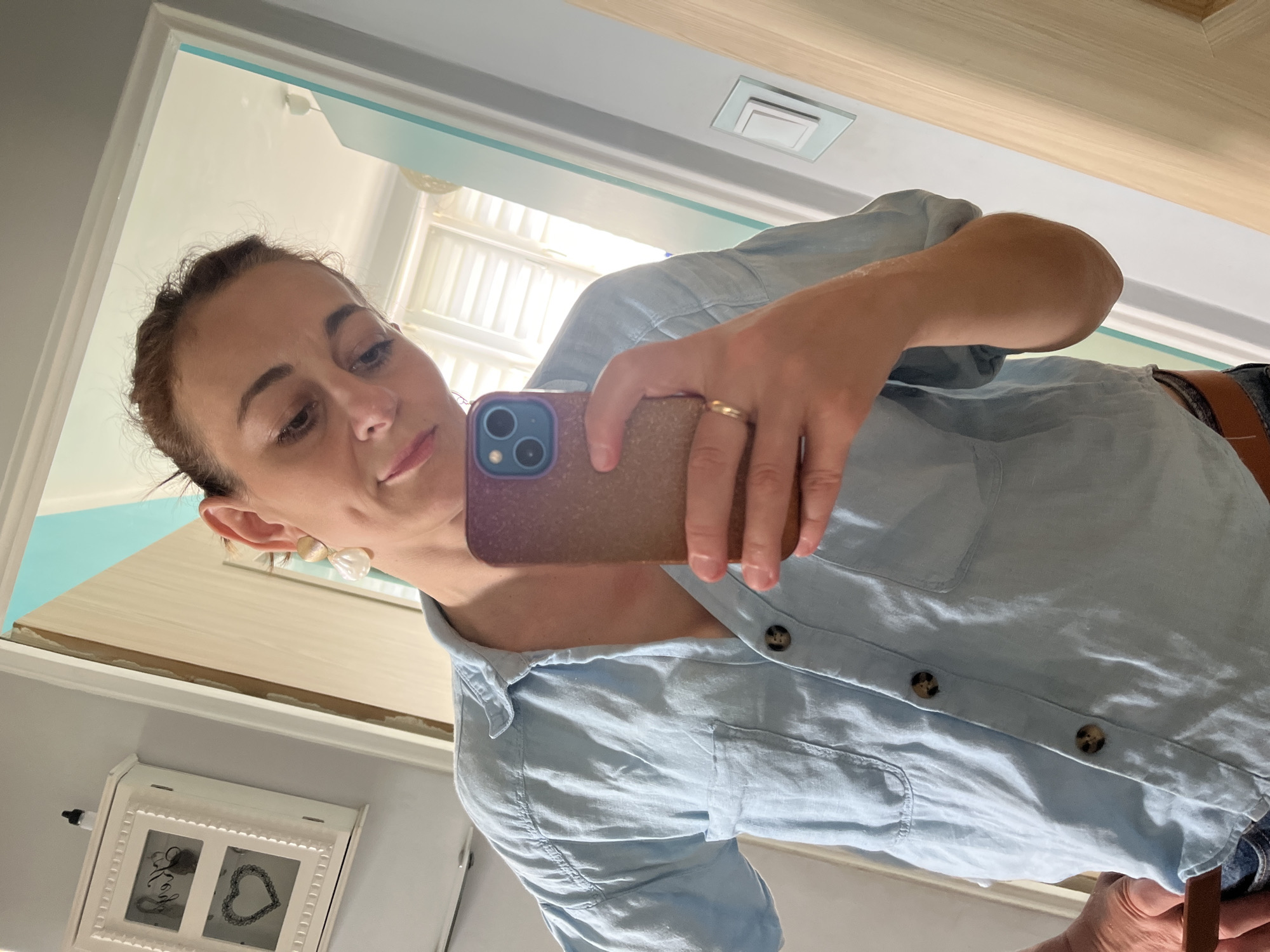 A woman is taking a mirror selfie in a bathroom. She is wearing a light blue button-down shirt. The bathroom has white walls and a wooden door. There is a small white cabinet on the wall with two windows, each with a heart design visible behind the glass. The woman is holding a phone with a purple case and wearing a gold ring on her left hand.