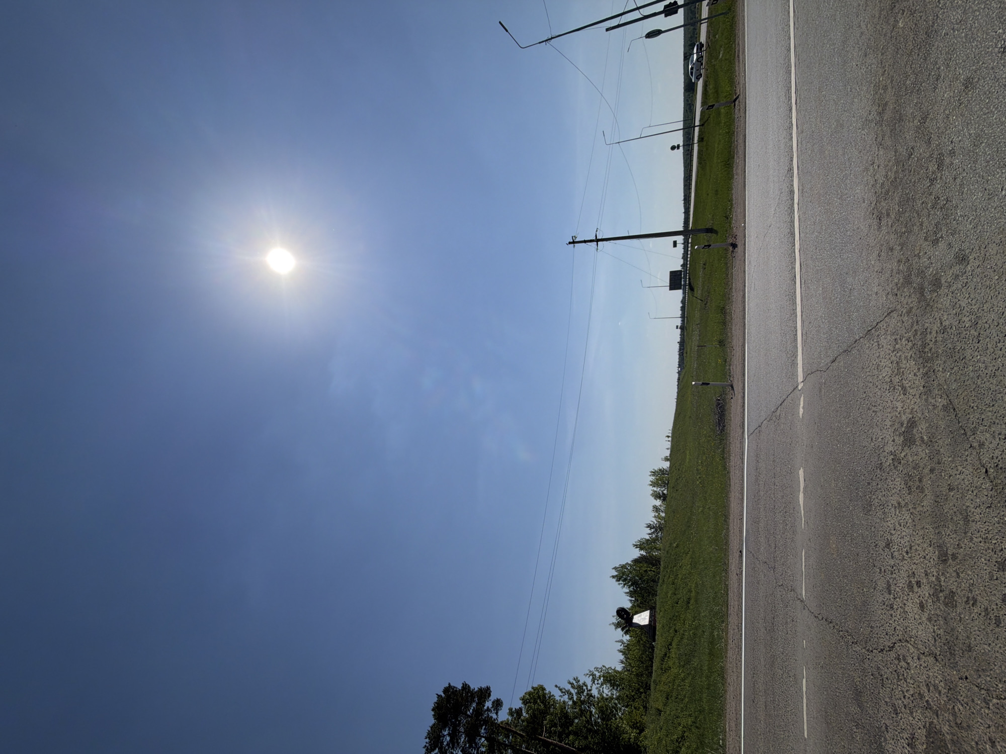 The image shows a sunny day near a road in Shaberdino, Russia. The sky is clear blue with the sun visible. A grey asphalt road stretches along the right side of the image, marked with white lane dividers. To the left of the road is a grassy verge with trees. Power lines and utility poles run along the edge of the road. In the distance, more trees and a vehicle can be seen.