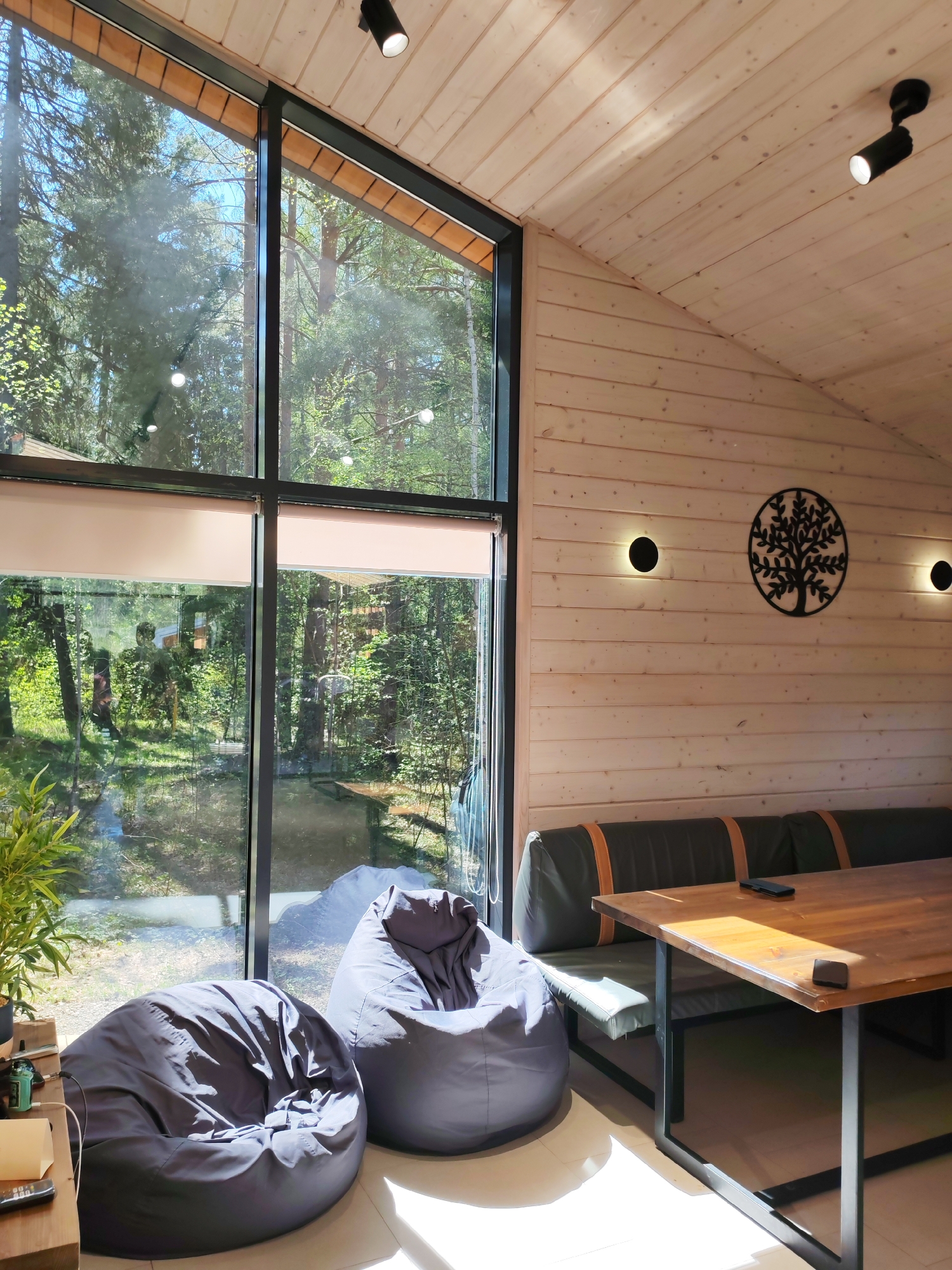 The image shows the interior of a room in a wooden house, possibly located in a wooded area near Izhevsk, Russia, based on the surrounding foliage visible through the large window. The room features light-colored wooden walls and a high, angled wooden ceiling. Natural light floods the room through a large window divided into sections by black frames. Outside the window, dense trees and greenery are visible, suggesting a forest or park setting.
Inside, there are two gray beanbag chairs near the window, positioned to capture the natural light. Next to one of the beanbags is a small table or shelf with some items on it, including what appears to be a plant.
Adjacent to the wall is a built-in seating area with a green cushioned bench and a wooden table with black metal legs. There are decorative wall lights and a metal tree design. The overall atmosphere is modern, cozy, and nature-oriented.