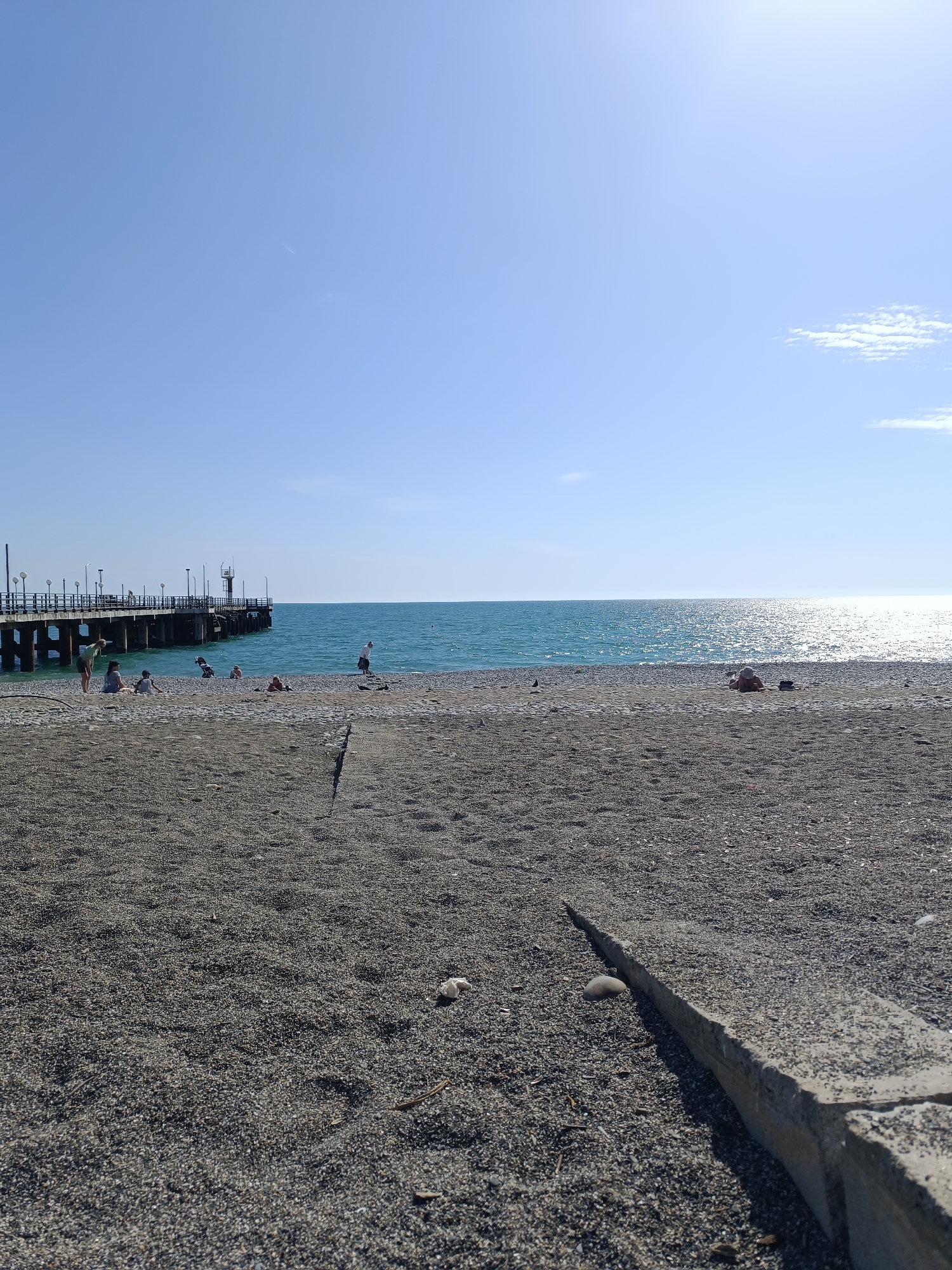 The image shows a beach in Unknown, Unknown. The beach is composed of gray pebbles and sand. A concrete barrier runs along the lower right of the frame. In the distance, there is a pier extending into the ocean. Several people are gathered near the pier. The ocean is turquoise in color, reflecting the sunlight. The sky is blue with a few scattered clouds.