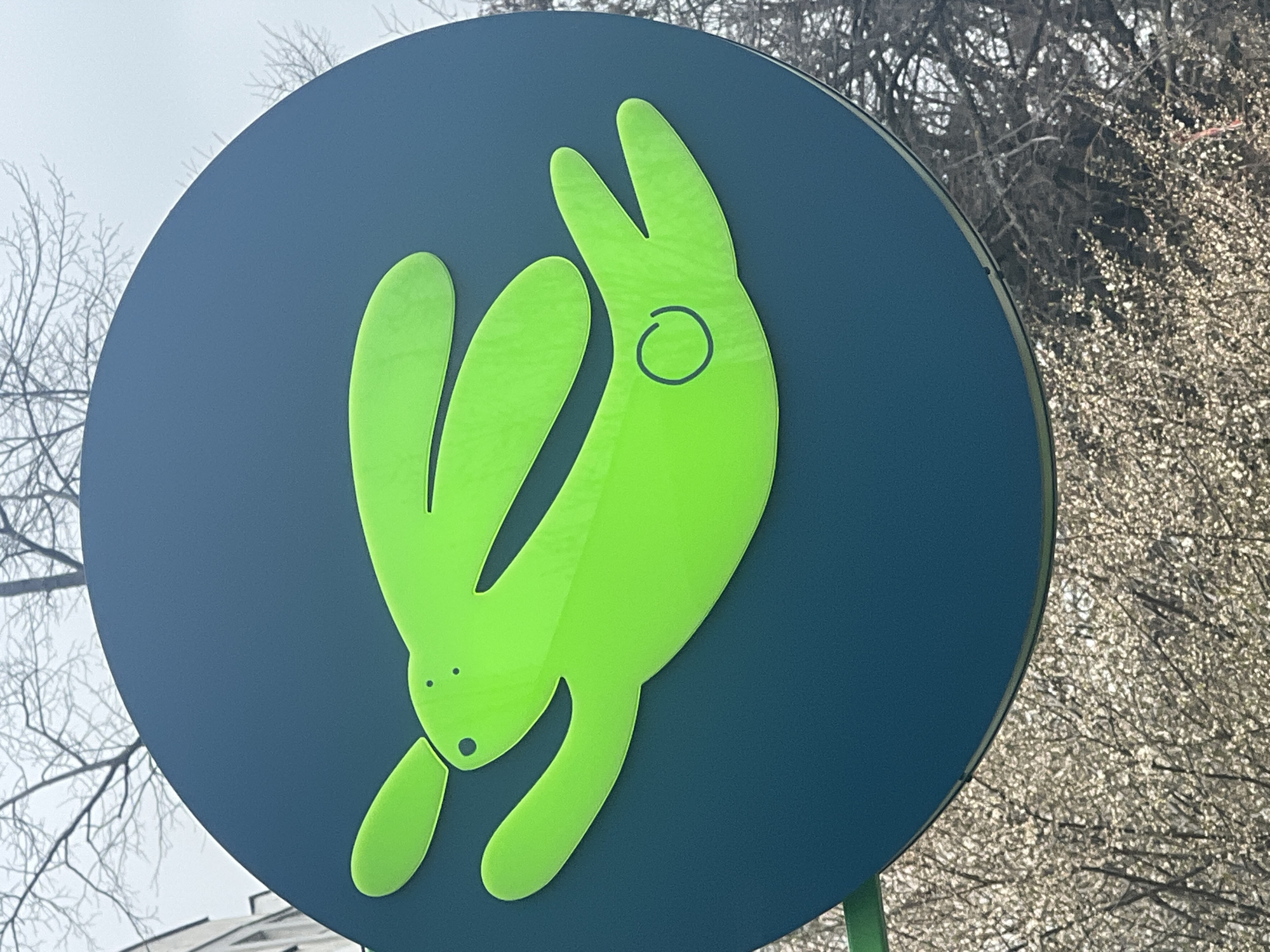 The image is a close-up of a sign in Sofia, Bulgaria. The sign is circular with a dark blue background. A stylized bright green rabbit figure is centered on the sign, oriented vertically with its head pointing downwards. The rabbit has long ears and a simple facial design with two small dots for eyes and a small oval for the mouth. A small circle is present on the rabbit's body. Bare trees are visible in the background.