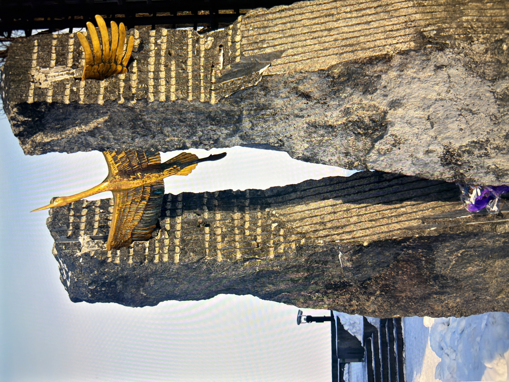 The image shows a monument in Kyiv, Ukraine, featuring a large, golden bird sculpture with outstretched wings mounted on two monolithic stone structures. The bird appears to be a crane or a similar large bird with detailed feathering. The stone structures are rough-hewn and textured, with vertical grooves carved into their surfaces. The monument is situated outdoors, with snow visible on the ground, suggesting a winter setting. In the background, there are hints of trees and a lamppost. Stairs and a walkway are visible to the left, also covered in snow. There is a small cluster of purple and silver items, possibly flowers or decorations, at the base of the right-hand stone structure.