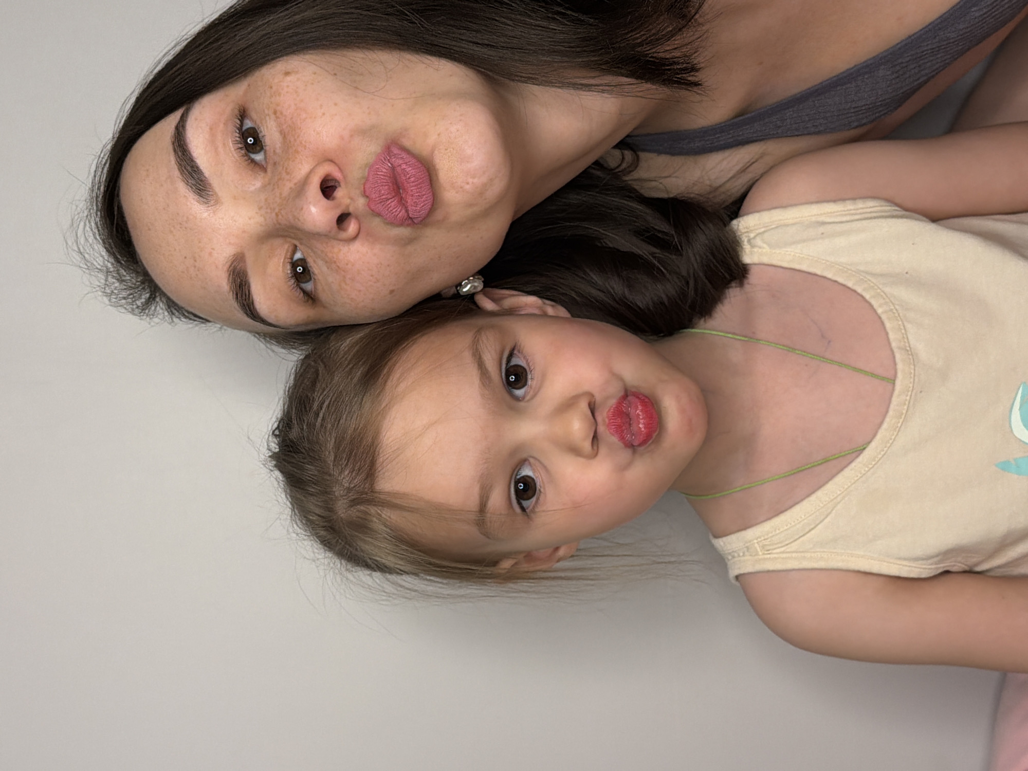 The image shows a woman and a young girl lying down side-by-side, posing with puckered lips. The woman has dark hair, freckles, and is wearing a gray top. The girl has light brown hair and is wearing a light yellow tank top. The background is a plain, light-colored wall. The location in the city of Chaikovsky, Russia cannot be determined from the image.