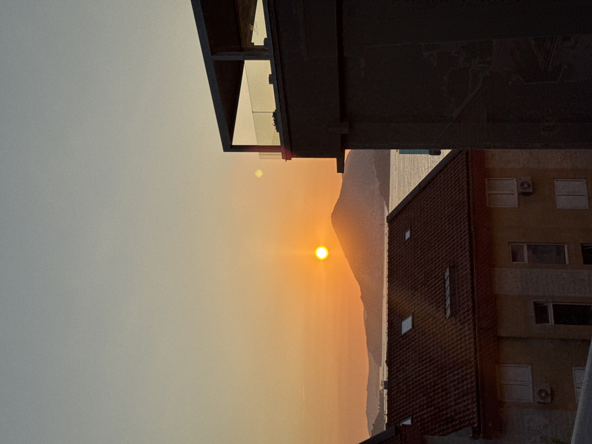 The image captures a serene sunset over the waters of Podličak, Montenegro. The sun, a bright orb of orange, is low on the horizon, casting a warm glow across the sky. Silhouetted against the vibrant sky are rolling hills and distant islands. In the foreground, the tiled roofs of buildings are visible, with one building on the right featuring a modern, dark-colored structure with a glass-railed balcony. The other building on the left is lighter in color, with visible windows and an air conditioning unit. The overall scene evokes a peaceful end to the day in this coastal Montenegrin town.