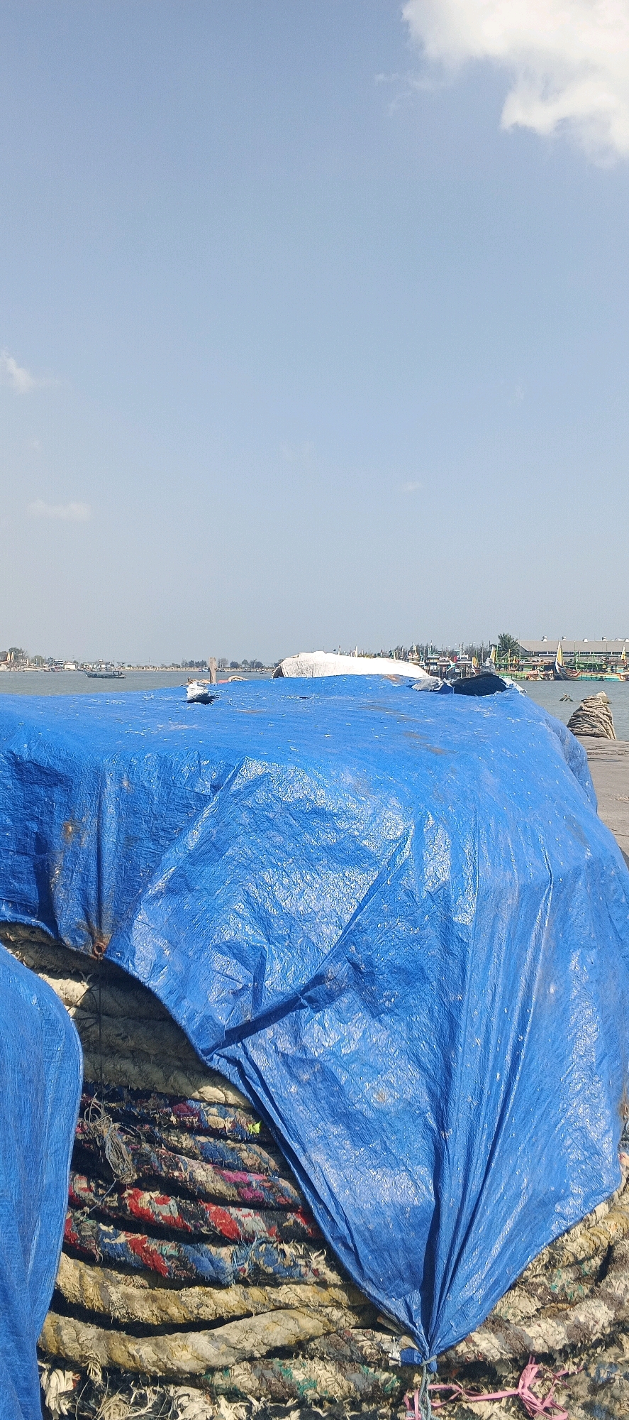The image shows a close-up of a large, blue tarp covering a large bundle of what appears to be old fishing nets or ropes, piled high. The material underneath the tarp is a mix of coarse fibers in various muted colors, including beige, brown, red, blue, and green. The tarp is wrinkled and has some discoloration, indicating it has been exposed to the elements. Part of another blue tarp is visible on the left side of the image. In the background, a body of water can be seen, with a distant shoreline populated by buildings and trees. Several boats are visible on the water and docked along the shore, suggesting a coastal or riverside location. The sky is clear and blue with a few scattered white clouds. The overall scene appears to be outdoors, possibly in a port or storage area. The specific location is identified as Blimbing, Indonesia, likely a coastal area given the presence of boats and water.