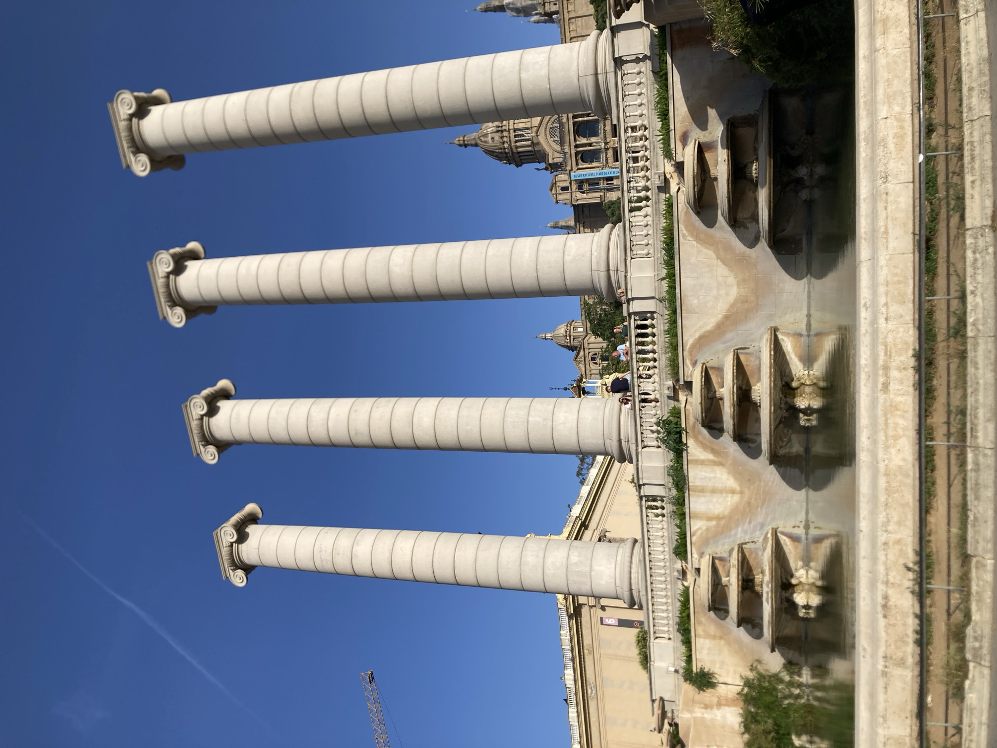 This is a photograph of the Palau Nacional in Barcelona, Spain, taken on a clear, sunny day. In the foreground, four large, white Doric columns stand tall against a bright blue sky. A contrail is visible in the sky above the columns. Behind the columns, a wide staircase leads up to the Palau Nacional, which is a grand building with domes and an ornate facade. The building is framed by trees and a balustrade. In front of the staircase, a tiered fountain with several basins is visible, with water flowing into a pool. The water reflects the surrounding architecture. There are a few people visible on the staircase and near the fountain. The scene is peaceful and grand, with the sunlight casting shadows and highlighting the architectural details.