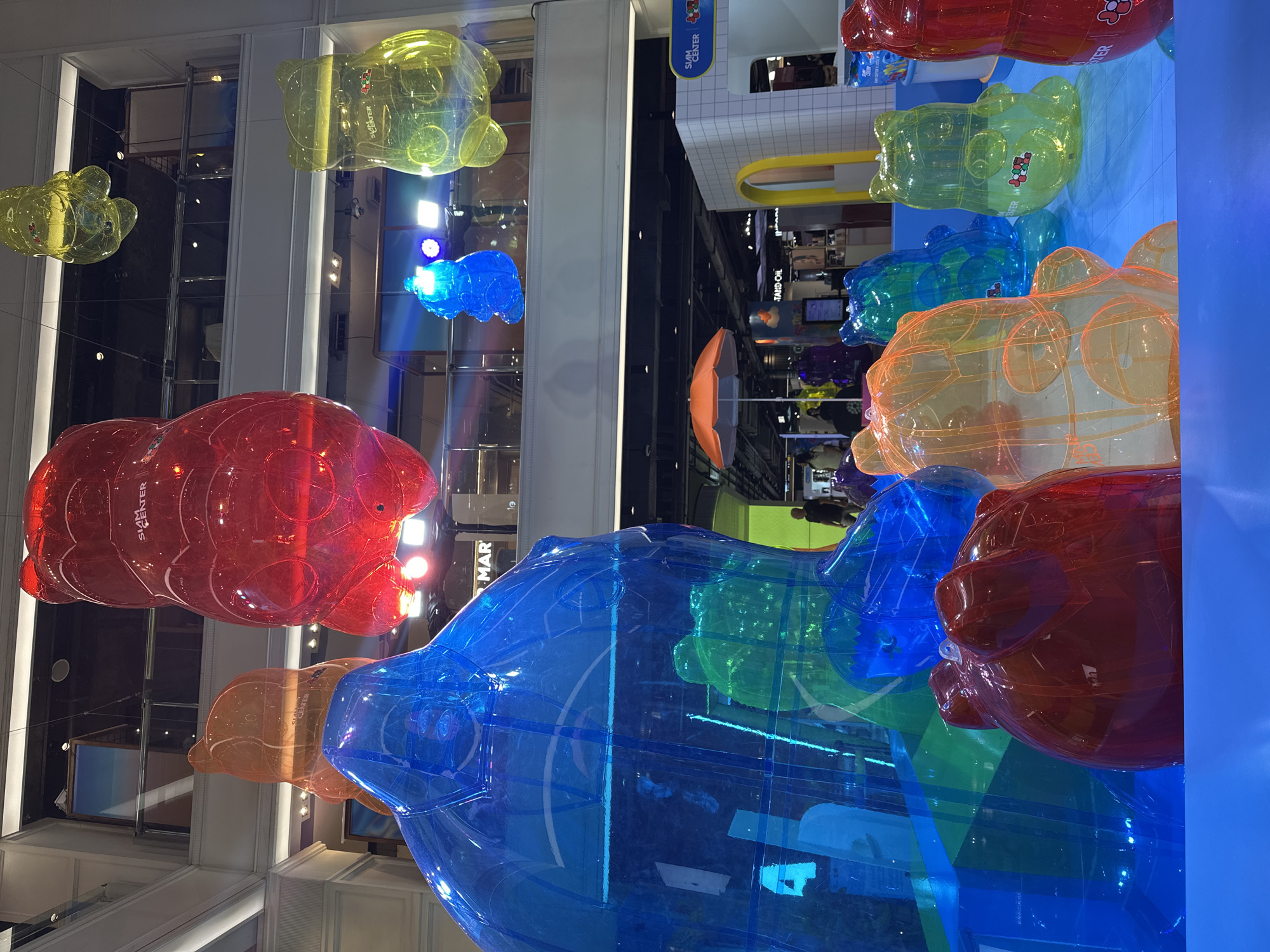 The image shows an indoor display of large, translucent, colorful bear-shaped inflatables. The bears are red, blue, yellow, and orange. The location appears to be a shopping mall or commercial space, based on the store fronts and architecture in the background. The location is likely in Bangkok, Thailand, based on the writing on the inflatables and the building's architecture. There are other people visible in the background.