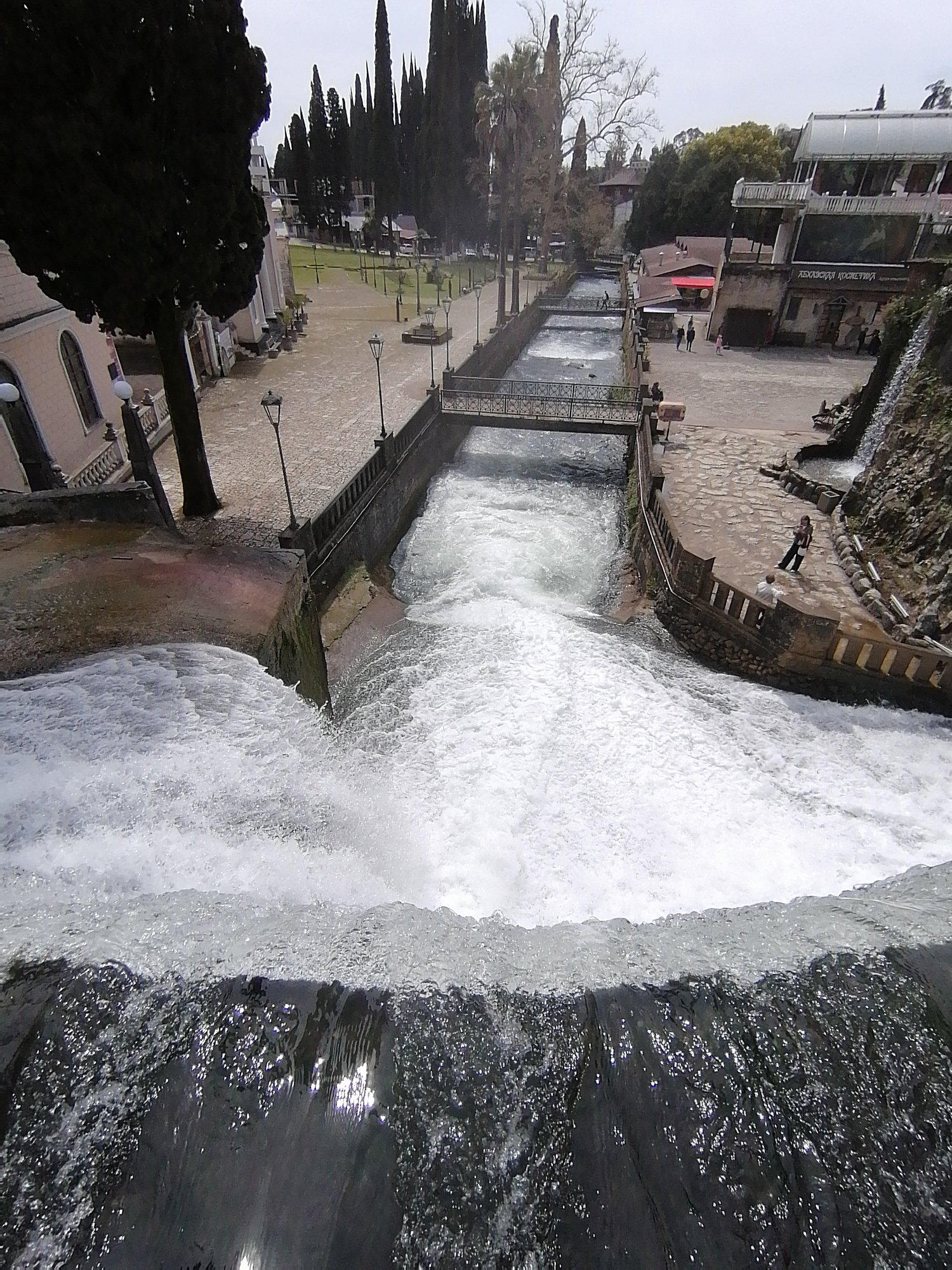 The image captures a wide-angle, high-angle view of a scenic urban or park environment centered around a flowing waterway and waterfall. In the immediate foreground, a powerful cascade of white, foamy water tumbles downwards, dominating the lower half of the frame. The water is highly turbulent, reflecting some light from the overcast sky.
Beyond this waterfall, a channelized river or canal flows, contained by concrete banks. Two pedestrian bridges with ornate metal railings span this waterway in the midground. Along the riverbanks and paved areas, several tall, dark coniferous trees, resembling cypresses, are prominent, creating a structured landscape. Scattered lampposts line the paths and river edges.
To the left, a light-colored building with classical architectural elements, including arched windows and balustrades, is partially visible, nestled amongst trees. On the right side of the canal, a multi-story building with modern glass sections and a roof structure is prominent. Directly below this building, a smaller, rocky waterfall feeds into the main canal. Visible text on the building reads "АБХАЗСКАЯ КОСМЕТИКА" (Abkhazian Cosmetics).
Several individuals are present in the scene. On the paved area to the right, near the "Abkhazian Cosmetics" building, a few people are standing, one in dark clothing and another in lighter attire. Further back, a solitary figure can be seen walking along a path next to the canal. Other indistinct figures are visible in the distance.
The setting appears to be a well-maintained public area, possibly a park or historical site, with paved walkways and green spaces. The sky is overcast, providing soft, diffused lighting with no strong shadows, suggesting daytime, possibly late morning or early afternoon. The trees in the background include both evergreens and some bare deciduous trees, hinting at late winter or early spring. The overall atmosphere is tranquil despite the powerful flow of water.