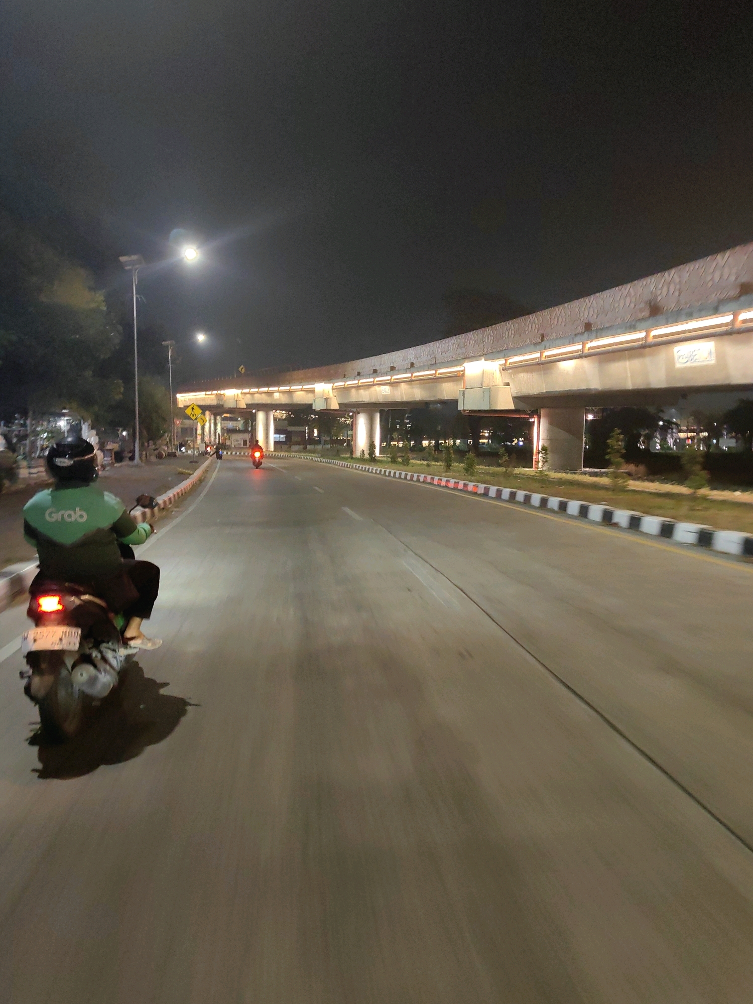 In Sawotratap, Indonesia, at night, a person wearing a green Grab jacket rides a scooter down a paved road. The road is lined with a curb that alternates between black and white squares, and there are lights lining an elevated overpass above the road. The sky is dark, with streetlights visible in the distance. The scooter has a license plate and its taillight is illuminated.