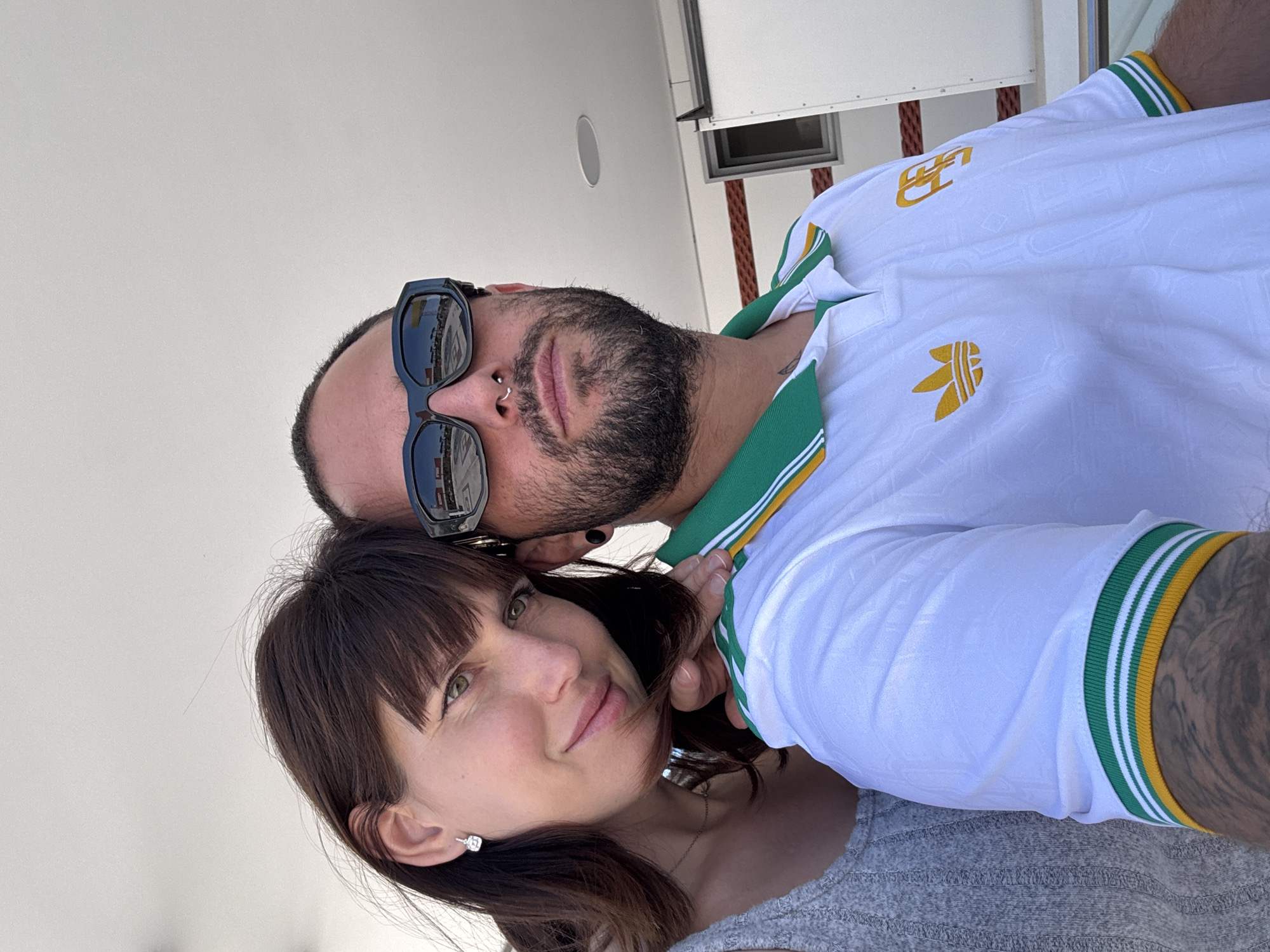 A selfie depicts a man and a woman positioned close together, possibly on a boat, though the location is not explicitly specified as Pomezia, Italy. The woman is in the foreground, wearing a grey shirt and a delicate necklace, with short brown hair featuring bangs. She has light eyes and an earring on one ear. Behind her, a man with a beard, a nose ring, and sunglasses rests his face near hers. He is wearing a white soccer jersey with green and gold trim and an Adidas logo. His arm is visible, showing tattoos. The background consists of a white wall, likely a part of the boat's structure, with a small speaker and a rectangular window visible. The lighting appears to be bright and natural.