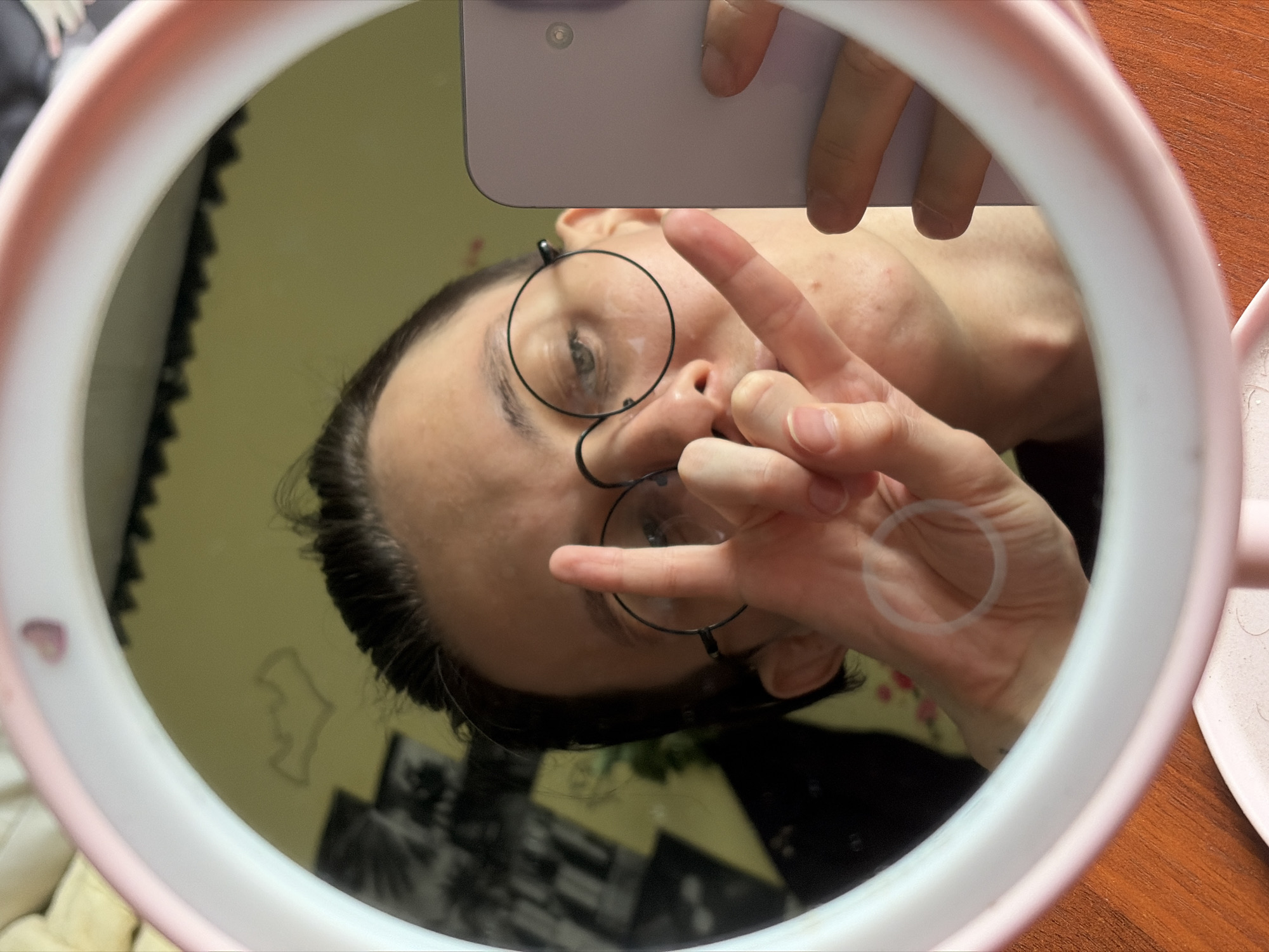 The image is a mirror selfie taken in what appears to be an indoor setting, possibly a room with a wooden surface and a light-colored wall in the background. The mirror is framed by a pink and white ring light.
The main subject is a person whose face is reflected in the mirror. They are wearing round-framed glasses and have dark hair pulled back. Their head is tilted, and they are holding up two fingers in a peace sign. Another hand is visible, also making a gesture. The person's expression seems neutral or slightly amused.
In the reflection, we can see parts of the person's upper body and their phone, which is being used to take the selfie. The background of the reflection shows a blurry environment with some light and dark areas, and potentially some objects. A faint outline of what looks like a building or a profile can be seen on the wall in the reflection.
The lighting is bright, likely from the ring light, illuminating the person's face. There is no indication of the time of day or weather conditions. The overall mood of the image is casual and playful, typical of a selfie. No visible text is present in the image.