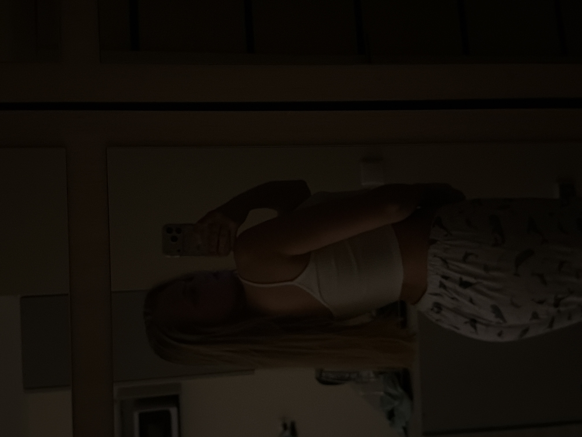 A woman with blonde hair, wearing a white tank top and patterned pants, is taking a selfie in a dimly lit room, reflected in a mirror. The environment shows a simple interior, with elements suggesting a bedroom or bathroom. The location is not discernible from the image.