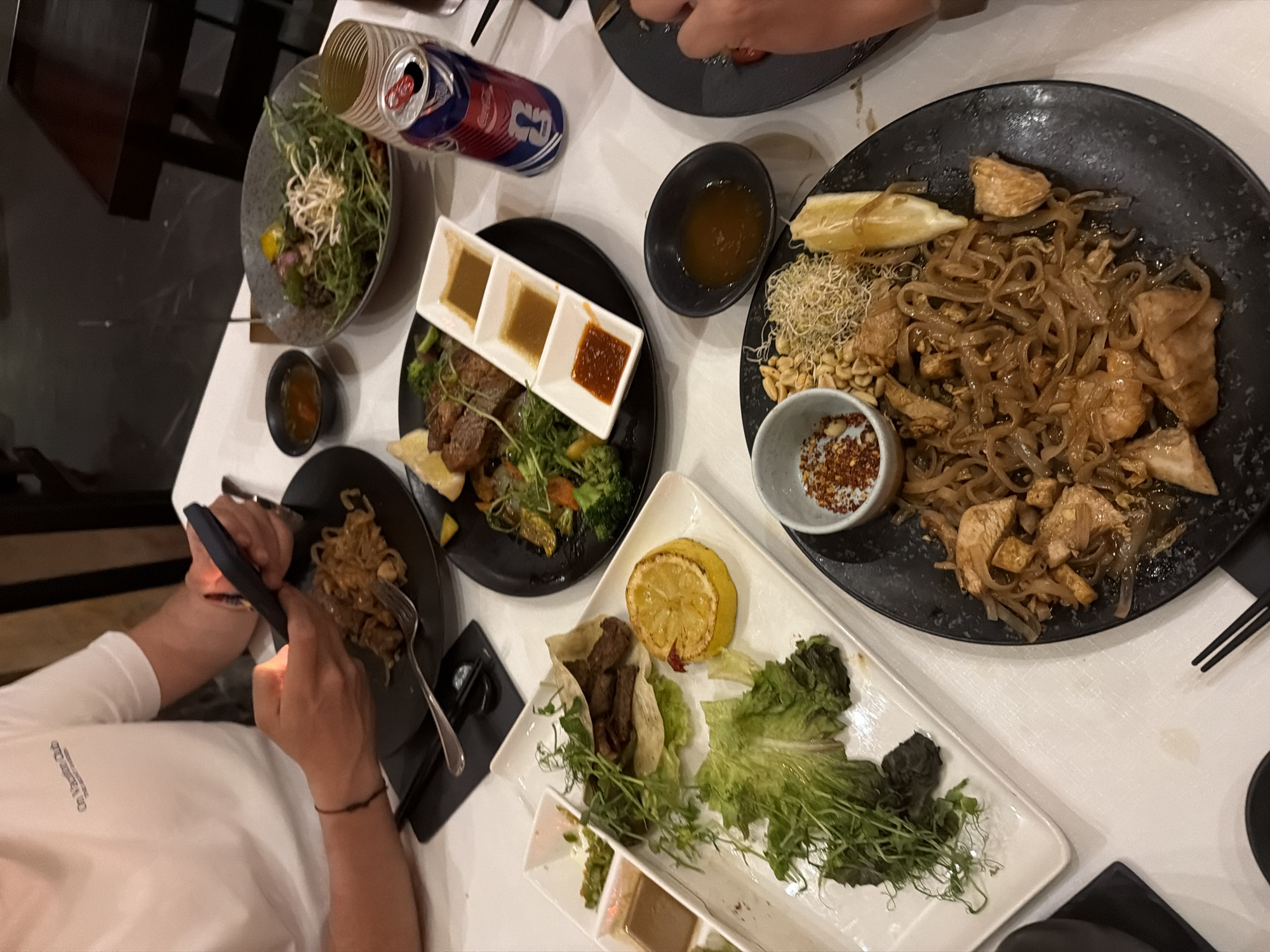 A tabletop scene shows an array of dishes, likely in a restaurant, in the city of Las Jarretaderas, Mexico. Various foods are presented, including a salad in a grey bowl, a black plate holding cubed steak and vegetables with a sauce sampler, a black dish containing Pad Thai with a lemon wedge and a small dish of chili flakes, and a white rectangular plate with lettuce, meat in a pita, lemon and a sauce sampler. A can of soda is also on the table. Two people are present. One is operating a mobile phone while holding chopsticks. The second person, located at the top of the photo, is possibly using silverware. The table has a white surface. Chopsticks are situated next to the Pad Thai plate. Other dishes include a black plate with an indiscernible food item and a black ramekin of what seems to be sauce.