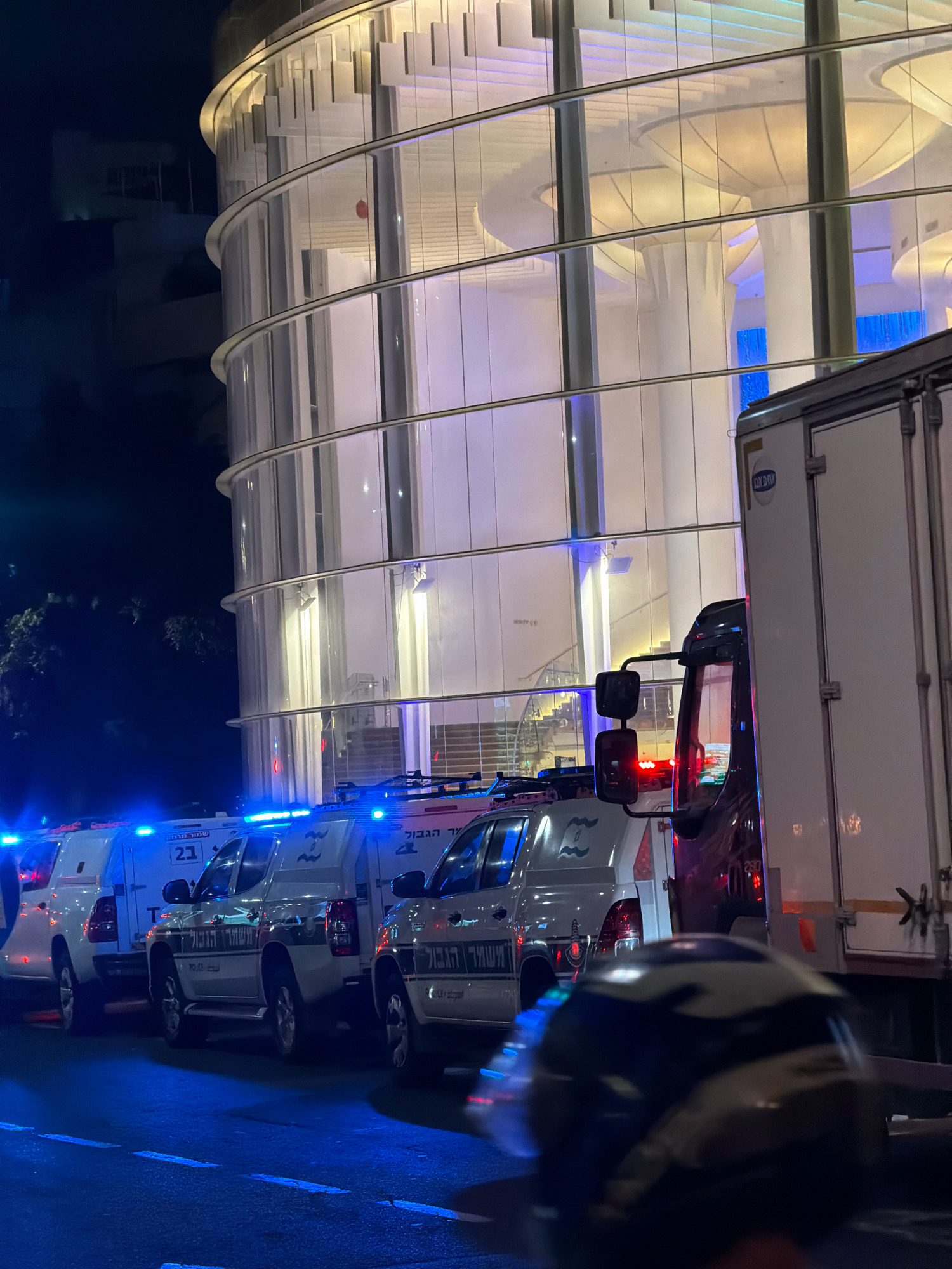 A night-time, eye-level, medium shot captures a row of white security or police vehicles parked on a street in Tel Aviv, Israel, in front of a modern, multi-story building with a curved glass facade. The vehicles, some with blue flashing lights, have Hebrew lettering on their sides, with one clearly showing the number "21". A large white truck with a closed cargo section is parked to the right of the security vehicles. The building's facade is illuminated from within by soft, warm light, creating reflections on the glass. The street in the foreground is dark with faint white lane markings. A blurred object, possibly a helmet, is visible in the immediate foreground. The overall scene suggests a security presence or an event taking place at the building.