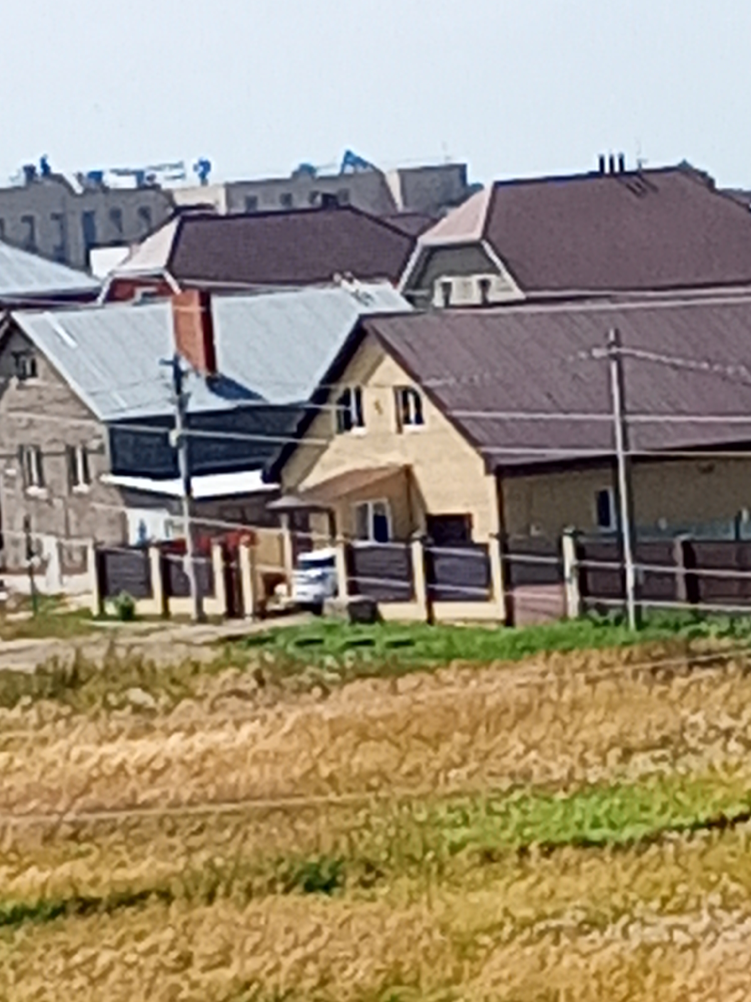 The image depicts a residential area in the city of Chistopol, Russia, characterized by several houses with visible rooftops and structures. In the foreground, a field of dry grass and some green vegetation extends towards the houses. The houses themselves exhibit diverse architectural styles, with pitched roofs of varying materials, including what appears to be metal roofing and shingles. One house is painted in a light yellow hue, while others have visible brickwork or siding. A white vehicle is parked near one of the houses. The scene appears to be bathed in daylight under a pale sky. The background shows more distant buildings, some of which appear to be under construction or renovation, with visible scaffolding and workers. The overall impression is one of a suburban or semi-rural environment undergoing development.