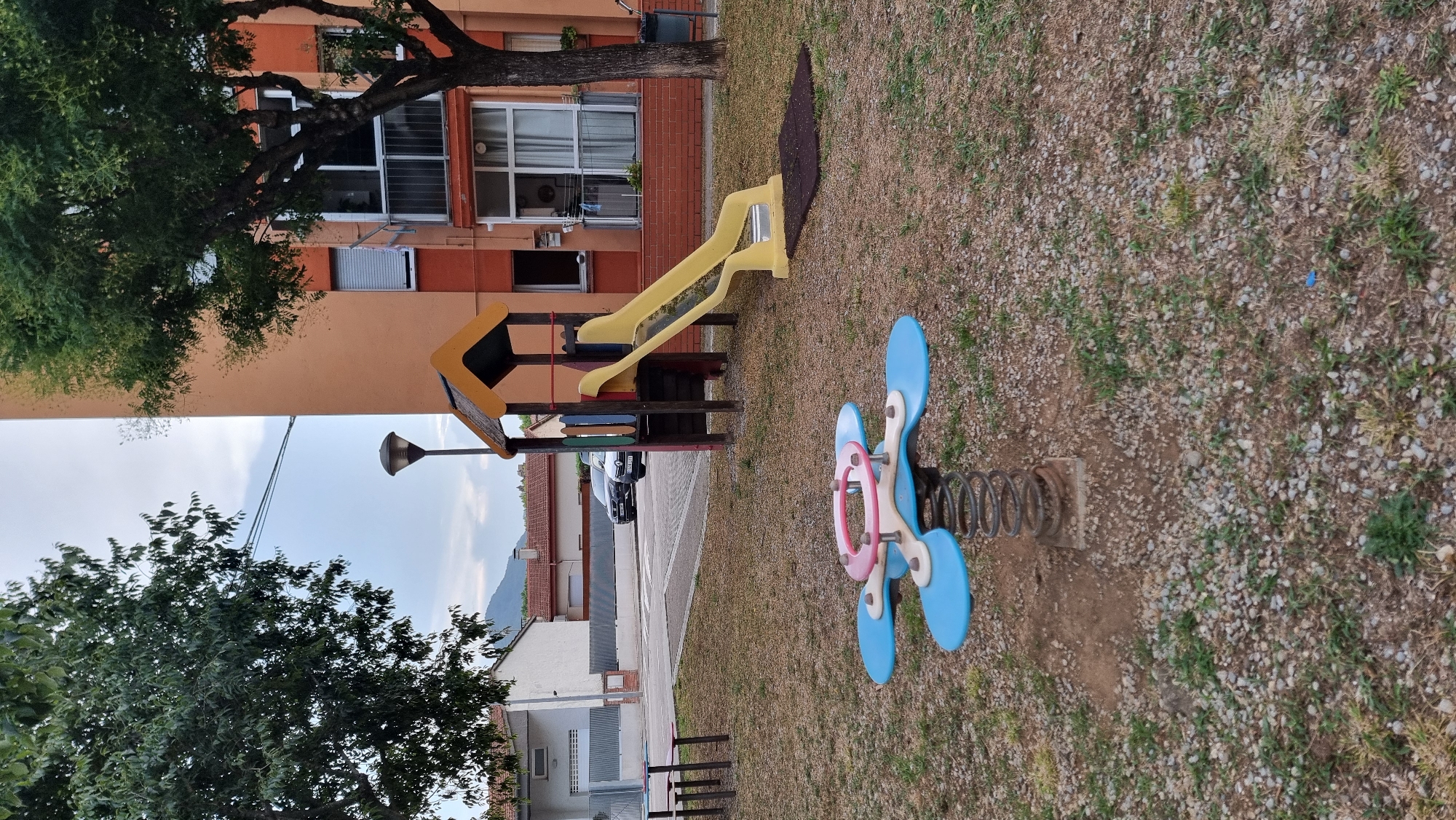 The image shows a children's playground with a yellow slide, a spring rider, and a picnic table in the foreground. The playground is located in the city of Sant Joan les Fonts, Spain, as indicated by the architectural style of the buildings and the surrounding vegetation. The playground is surrounded by gravel and dry grass. A large orange apartment building is visible in the background, as are some trees and a street with cars. The sky is cloudy.