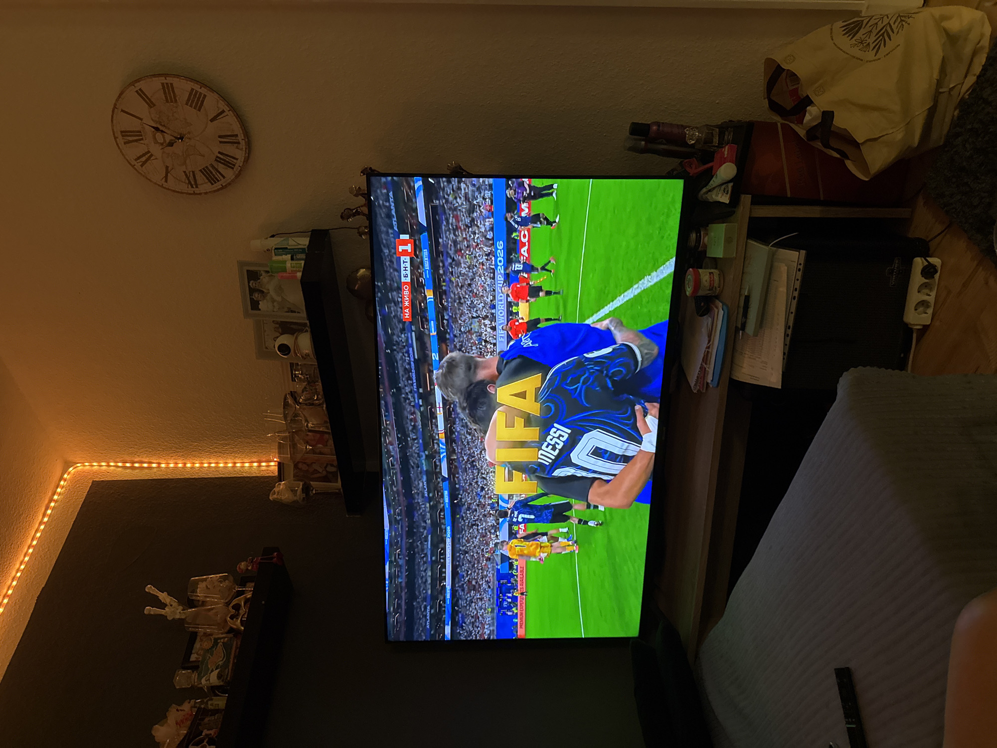The image depicts an indoor scene, likely a living room, where a large television is displaying a football match. The television screen shows a soccer game with players on a green field, and the word "FIFA" is prominently displayed. One player on the screen is wearing a blue jersey with the number 10 and the name "MESSI" visible.
The room is decorated with ambient lighting from string lights along the ceiling corner. On shelves above the TV, various decorative items and framed pictures are visible. A wall clock with Roman numerals hangs on the right side of the room. In the foreground, a bed covered with a grey textured blanket is partially visible, along with a remote control. A desk area is situated below the television, holding papers, a laptop, and some bottles. A large yellow shopping bag is on the floor next to the desk, and a power strip with plugs is visible near the floor.
The image's metadata does not contain information about its specific location, such as Lübeck, Germany. Therefore, the location in Lübeck, Germany cannot be confirmed or described based on the provided image.