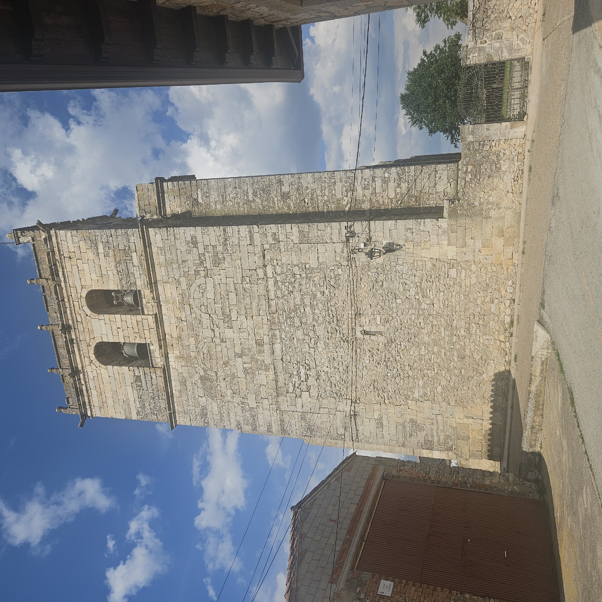 The image depicts a vertical, upward-angled view of a tall, rectangular stone structure in Algora, Spain. The structure appears to be a church tower or similar historical building, constructed from light-colored stone blocks. It features two arched openings near the top, likely containing bells. Ornate stone detailing adorns the edges and roofline.
The sky is visible behind the tower, displaying a bright blue hue with scattered white clouds. Electrical wires run across the view.
A building sits at the base of the tower. A nearby street is visible on the right side of the image. The building appears old and rustic, likely a part of the town's architecture.