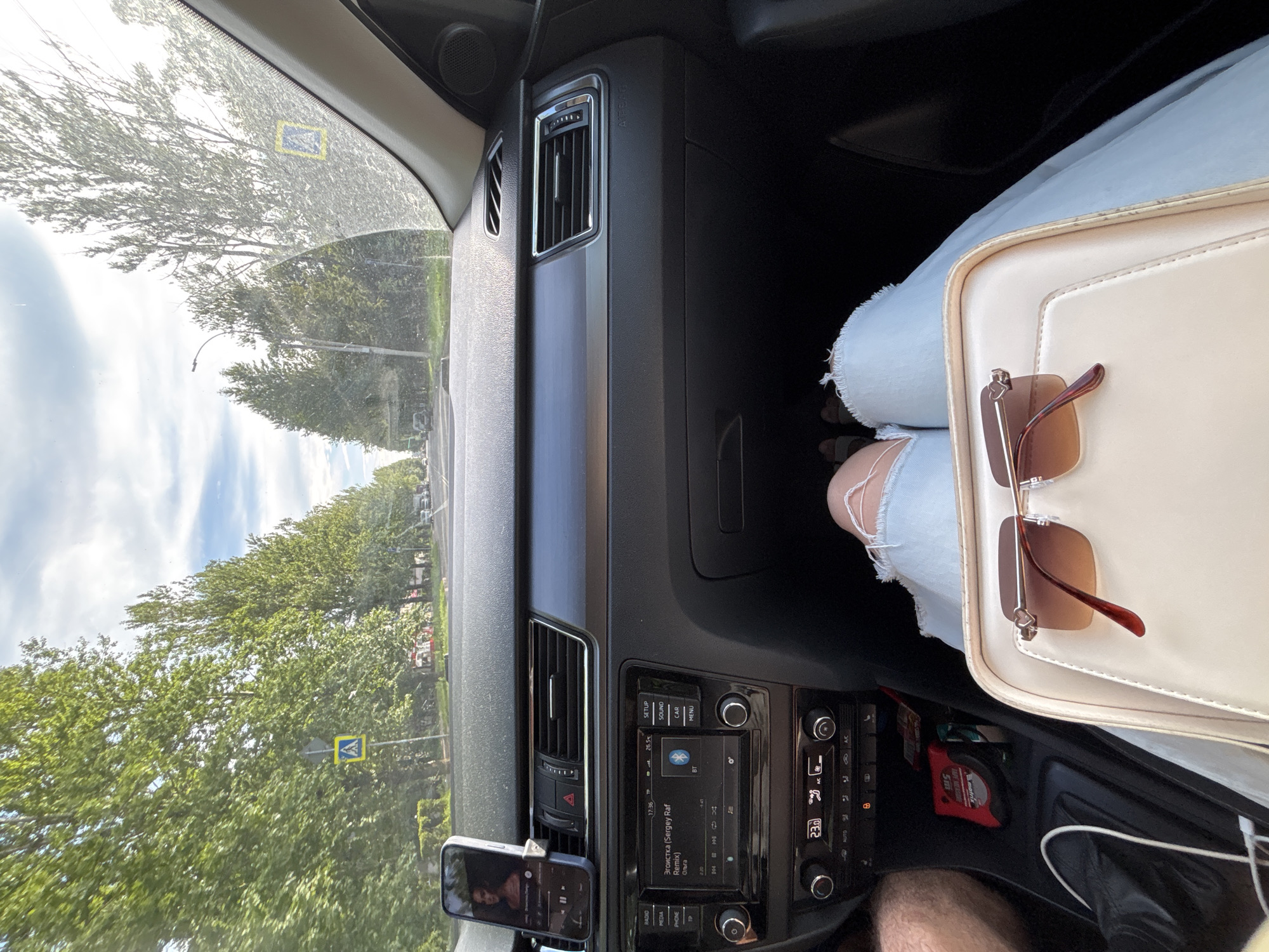 The image captures an interior view from the passenger seat of a car, looking forward. The car's dashboard, including the infotainment system displaying song information, air vents, and climate controls, is visible. A smartphone is mounted on the dashboard, playing music. Outside the car, a tree-lined street with a clear sky and clouds is visible. Pedestrian crossing signs are present on the side of the road. The car appears to be in motion. A person's legs, clad in ripped denim shorts, are visible in the foreground. A beige purse with sunglasses resting on it is also in the foreground. The location is specified as Rusanovo, Russia.