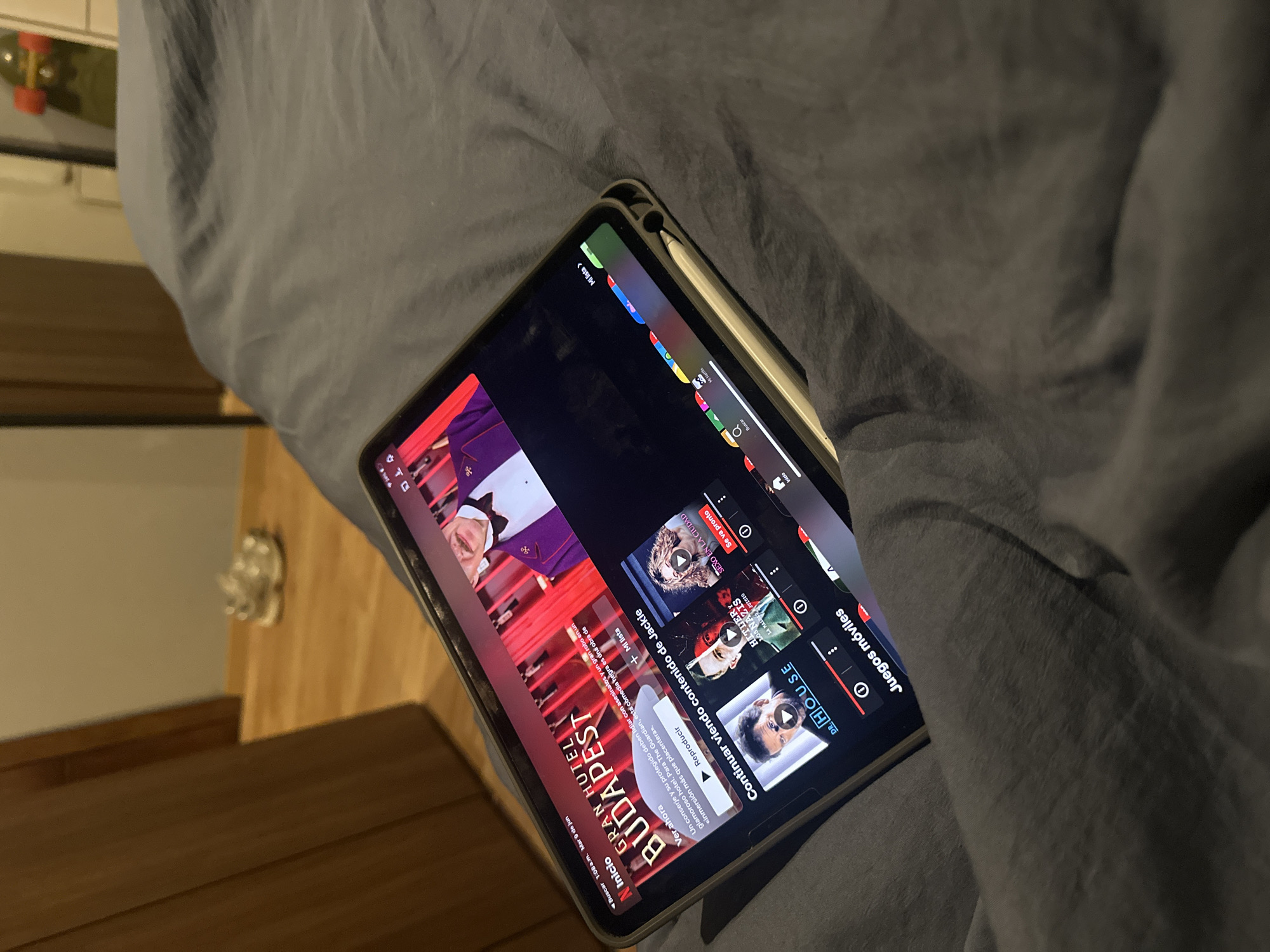 An iPad is propped on a gray, rumpled comforter, displaying a Netflix interface. The screen shows the movie "The Grand Budapest Hotel" as the primary content, with options to "Play" or "Continue watching content by Jackie." Below this, a row of smaller thumbnails includes "Dr. House," "Hitler: The Rise of Evil," and "Sex in the City." A stylus is positioned along the right edge of the iPad. In the background, a wooden door and wall are visible, along with a pair of white sneakers on a wooden floor. A skateboard with red wheels is propped against a wall in the far background. The scene appears to be indoors, likely in a bedroom. There is no indication of the location being in the city of Huixquilucan de Degollado, Mexico.