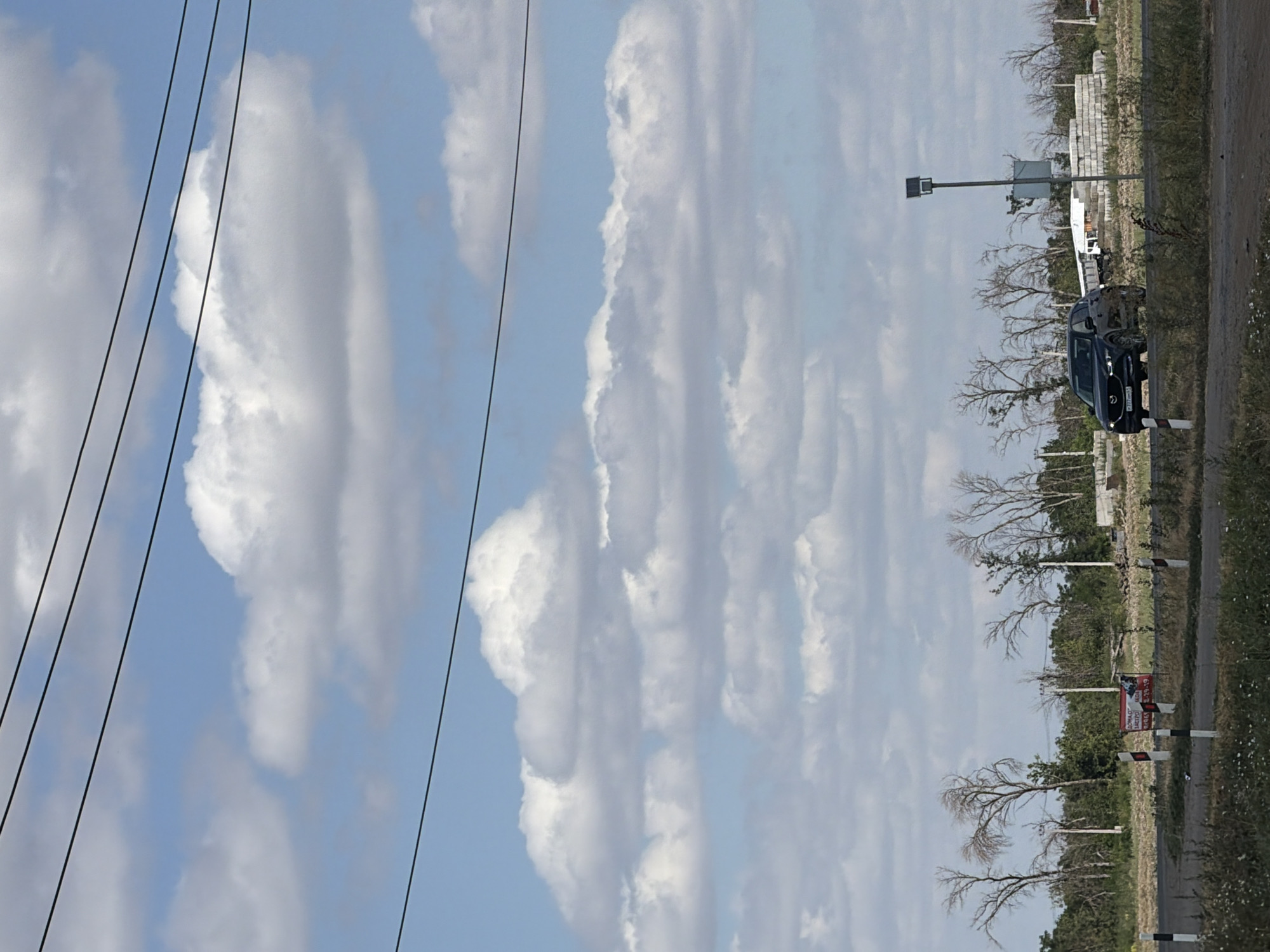 A dark-colored Mazda CX-5 is driving on a road in Krasnyi Krym, Russia. The sky is blue with scattered white clouds. There are power lines crossing the frame. In the background, there are several bare-branched trees, some bushes, and construction materials including concrete blocks and a white building. A sign is visible on the left side of the road.