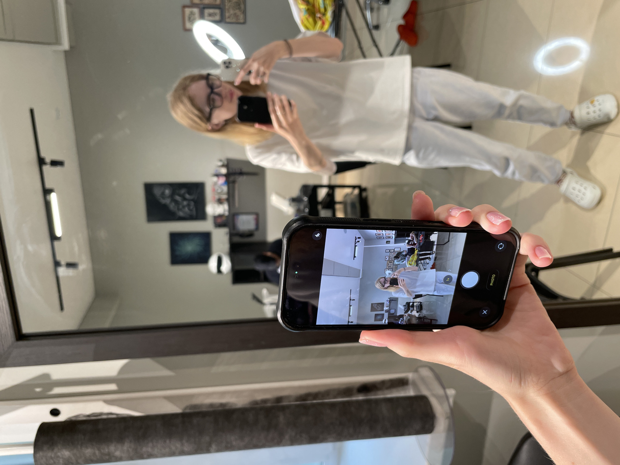 In a brightly lit room, a person is taking a selfie using a smartphone held in their left hand, which is visible in the foreground. The phone displays a reflection of the person taking the photo, who is standing in front of a large mirror. The person in the mirror is wearing glasses, a white t-shirt, and light-colored pants, and is holding another smartphone. The room appears to be a studio or salon, with various equipment visible in the background, including a ring light, shelves with items, and what looks like tattoo equipment. The overall environment suggests a professional setting. The image does not provide enough information to determine the specific location within the city of Izhevsk, Russia.
