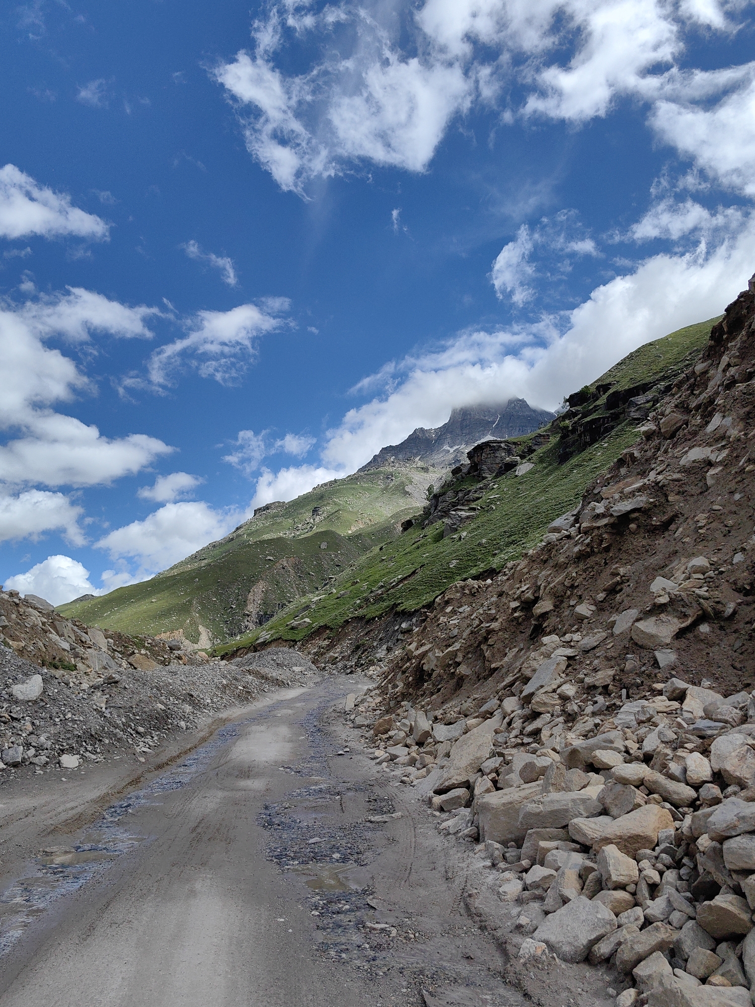 The image shows a narrow, unpaved road winding through a mountainous, rocky terrain. The road is wet and has puddles, suggesting recent rain. On either side of the road, there are steep slopes covered with loose rocks and stones, as well as patches of green grass. In the background, a large, green mountain rises, with its peak partially obscured by clouds. The sky is a vibrant blue, dotted with fluffy white clouds. The overall impression is one of a remote, rugged landscape, likely a mountain pass or trail in a rural area. Based on the context of Bashisht, India, this scene likely depicts a challenging road in the Himalayan region. There are no people or man-made structures visible in the immediate scene.