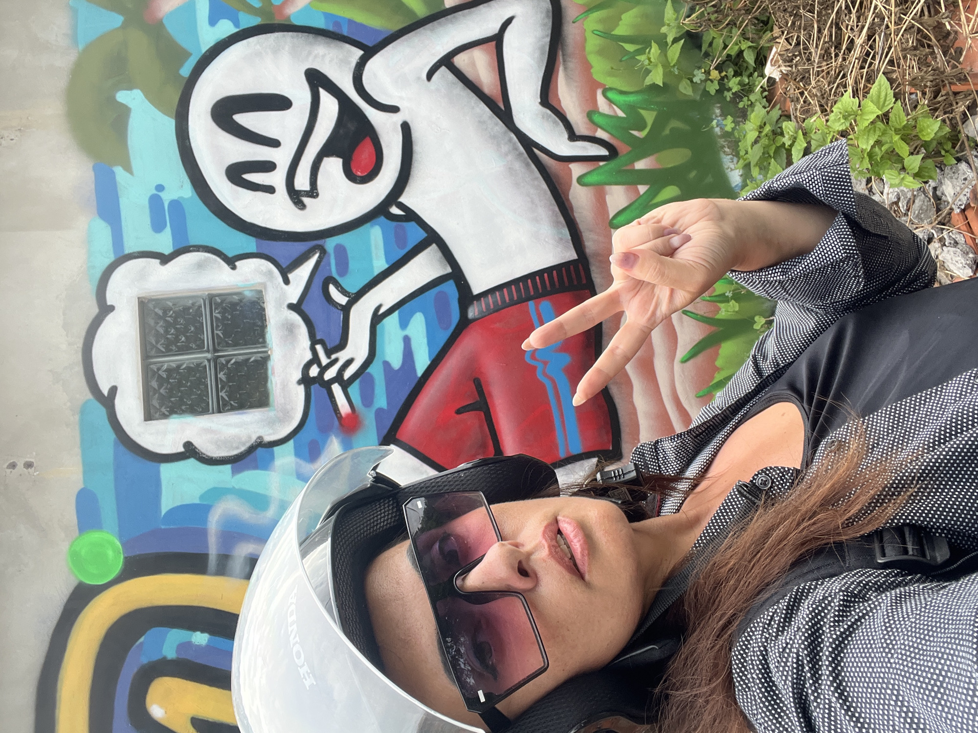 A woman wearing a white Honda helmet and large sunglasses is taking a selfie in front of a colorful graffiti mural. The mural depicts a cartoon character with a large smiling face, wearing red shorts, and holding a cigarette. The background of the mural features blue water, palm trees, and abstract yellow and black shapes. The woman is wearing a black tank top and a black and white patterned jacket. She is holding up two fingers in a peace sign. The setting appears to be outdoors, possibly a street in Kuta Selatan, Indonesia, with some greenery and rocks visible in the foreground.
