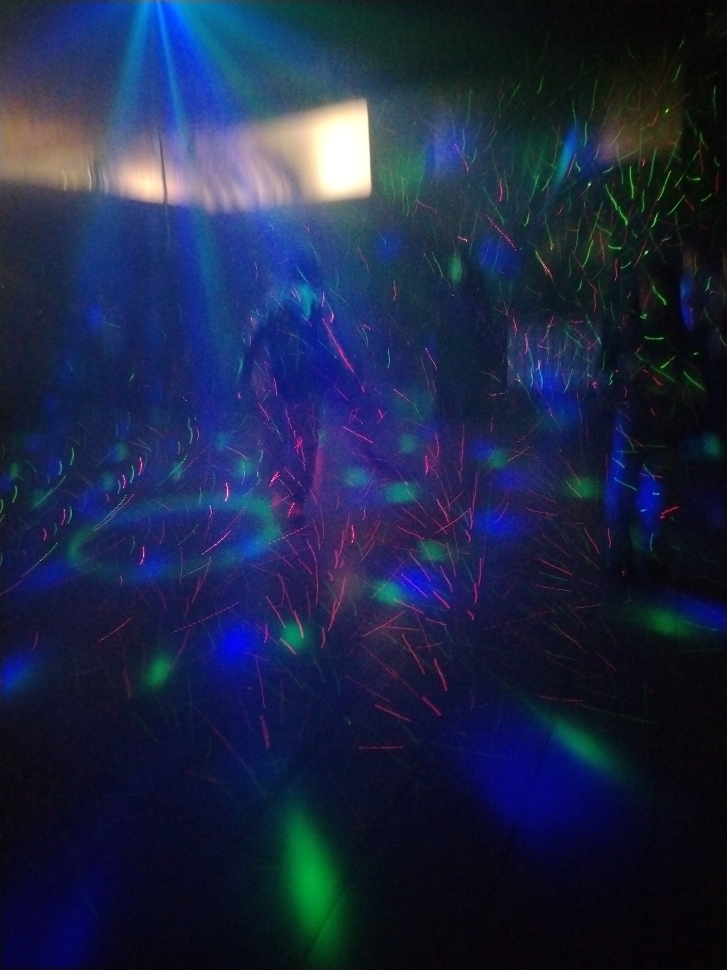 The image depicts an indoor scene illuminated by a combination of light sources including a rectangular white light and colored lights. These colored lights include green, blue and red. These lights are creating a light show with static lines of light that have a fuzzy look. The location, Rusanovo, Russia, is not apparent in the image. The image does not show any people, specific objects or actions.