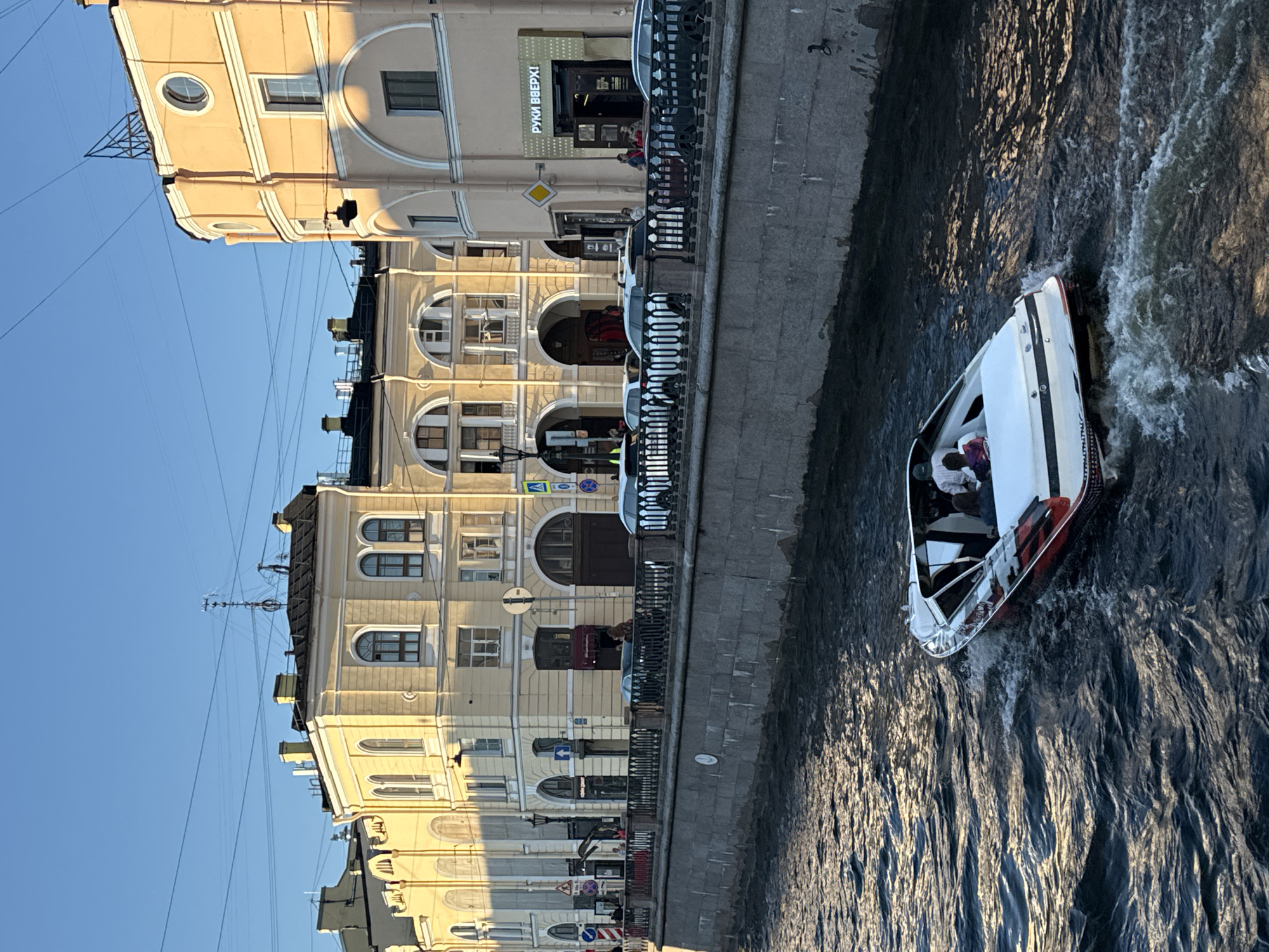 The image captures a view along a canal in Saint Petersburg, Russia. In the foreground, a white and red speedboat navigates the dark, rippling water, creating a wake as it moves. The boat carries at least three passengers, two of whom are visible in the rear.
Along the canal's edge, a stone embankment forms a barrier, topped by a black metal railing. Beyond the railing, a sidewalk runs parallel to the canal, with several parked cars visible. The architecture on the far side of the canal is characteristic of Saint Petersburg, featuring light yellow and peach-colored buildings with arched windows and classical detailing. One building has a sign with Russian text, "РУКИ ВВЕРХ!", which translates to "Hands Up!". The clear blue sky above is crisscrossed with power lines. The overall lighting suggests it is late afternoon or early evening, with the setting sun casting a warm glow on the buildings.
