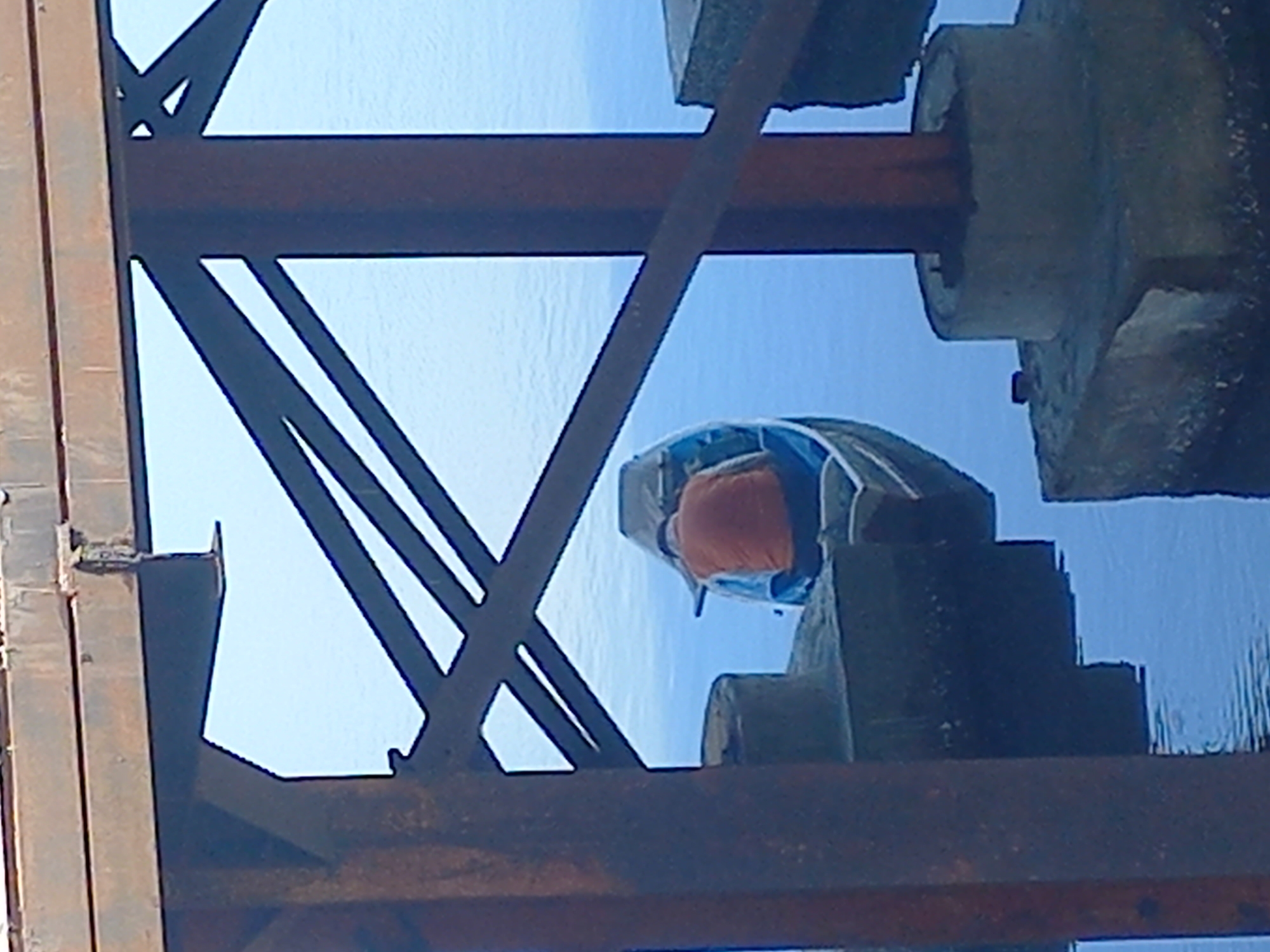 The image shows a person wearing an orange head covering and a light blue and grey protective suit standing on a concrete pier. The pier is located in Lhokseumawe, Indonesia, partially submerged in water. The view is obscured by a rusted metal structure with intersecting beams. The sky is visible in the background.